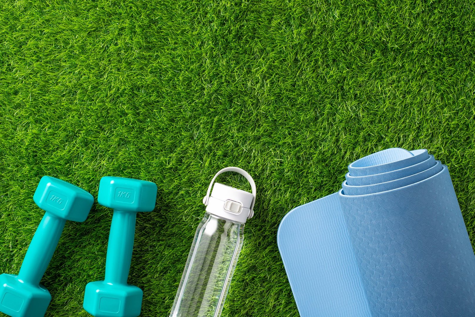 Fitness equipment including weights, a water bottle, and a yoga mat placed outdoors on green grass. Promotes a healthy, active lifestyle and represents exercise and wellness concepts