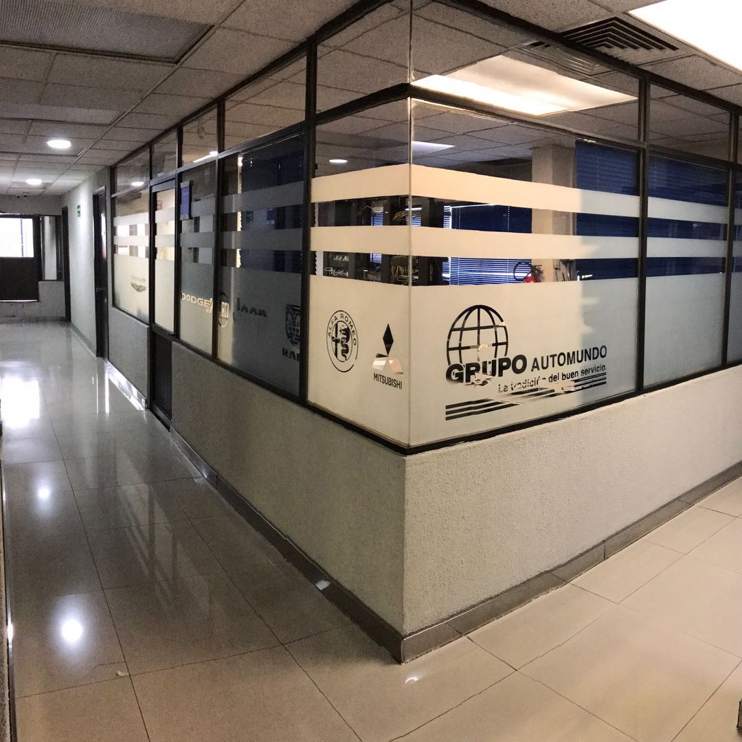 Office corridor with glass-walled partitions and company branding on the windows. Polished tile floor and ceiling lights.
