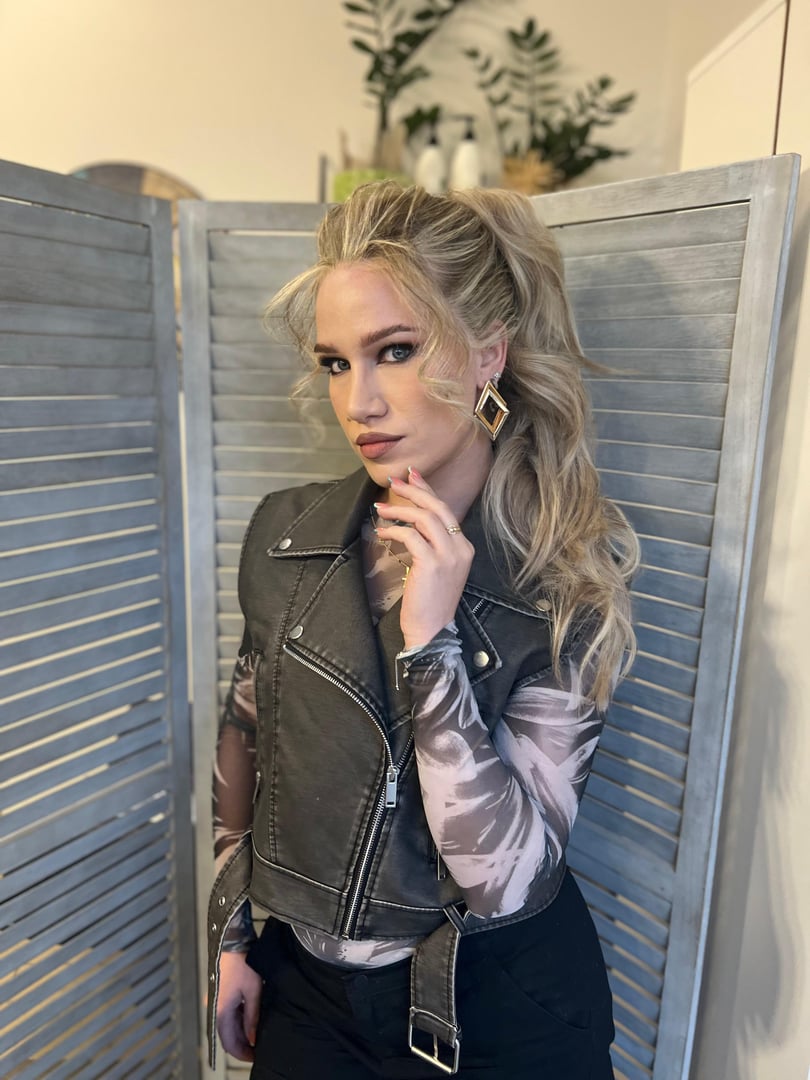Woman with blonde hair in ponytail wearing a black vest and earrings, standing in front of a wooden room divider.