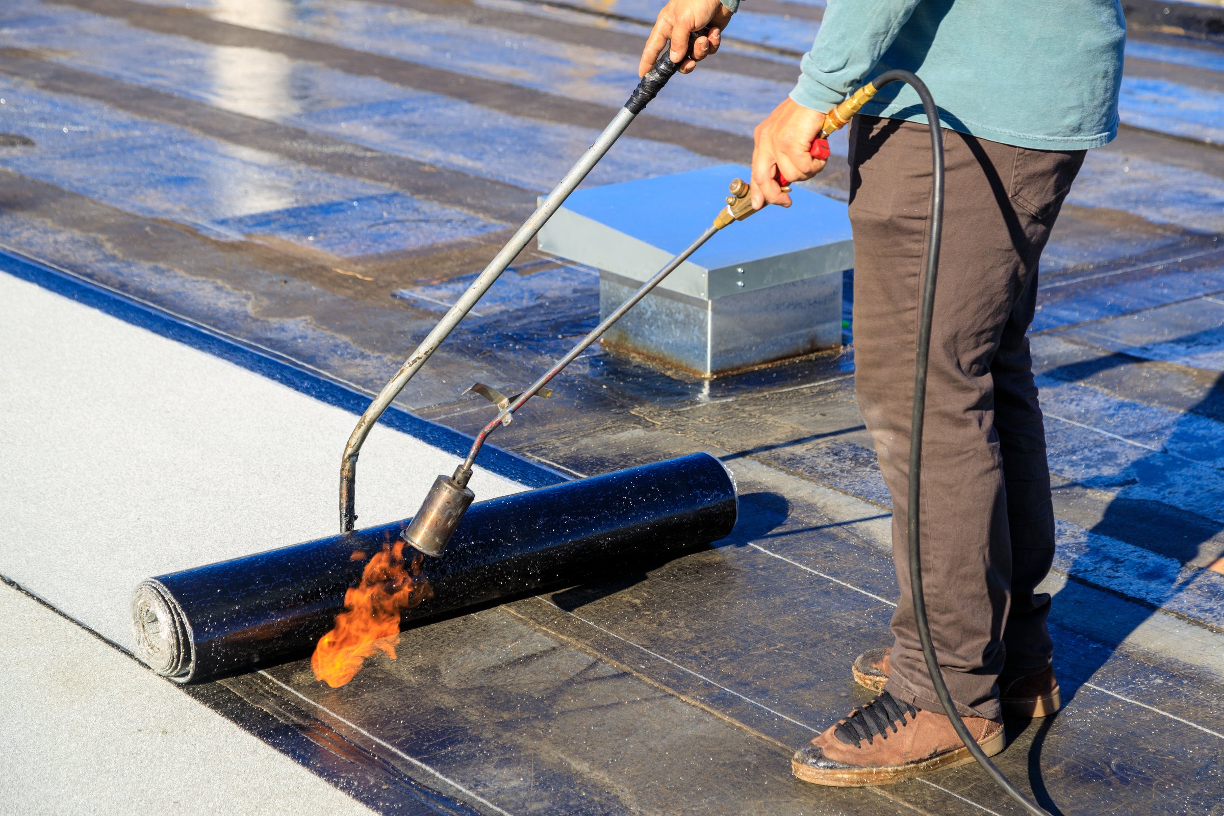 A roofer torches a membrane layer on a flat roof using a tiger torch to seal the asphalt bitumen, ensuring a durable waterproof layer.