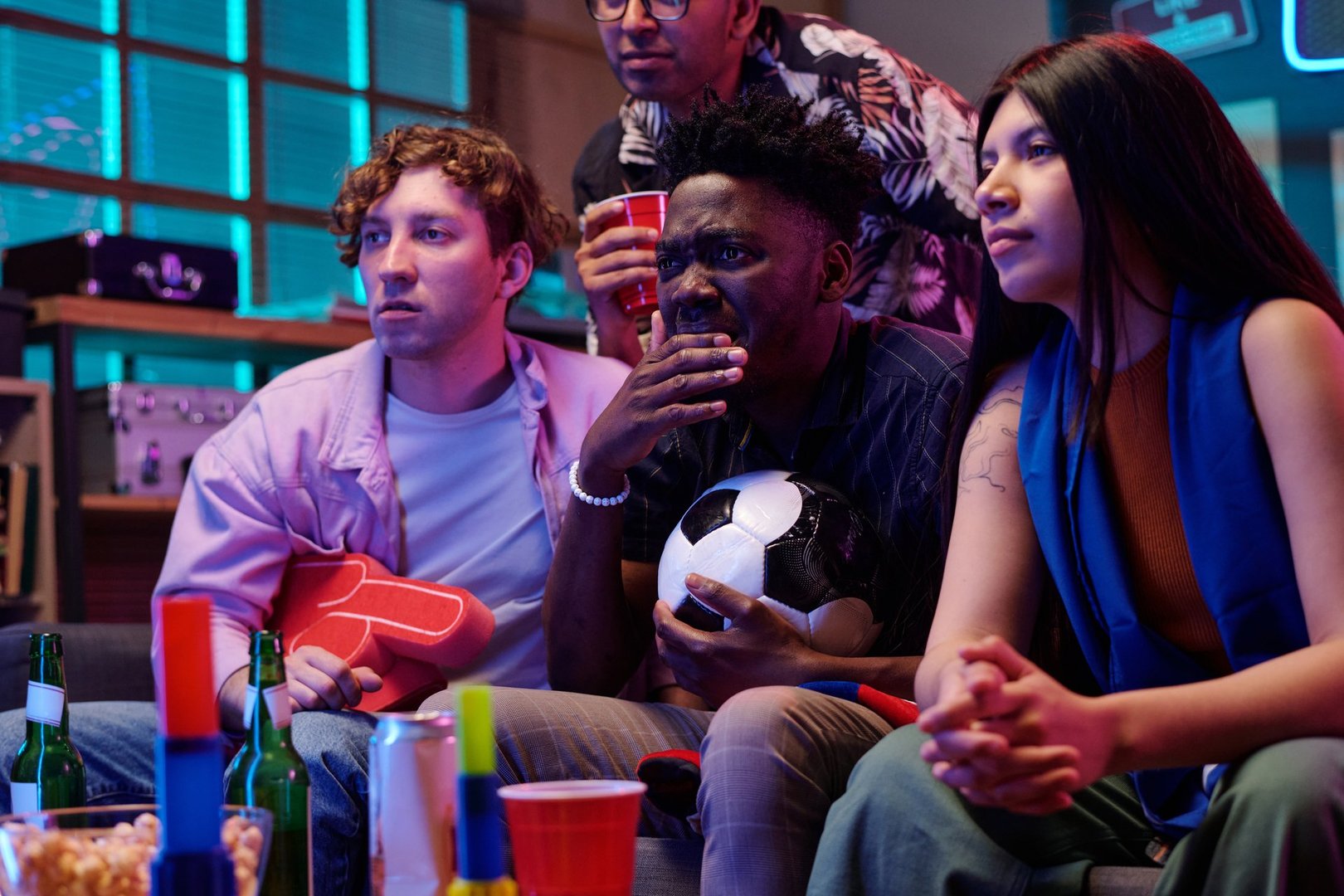 Multiethnic group of young adults watching sports game on television, Black man holding soccer ball with intense expression, friends sitting together with drinks and snacks