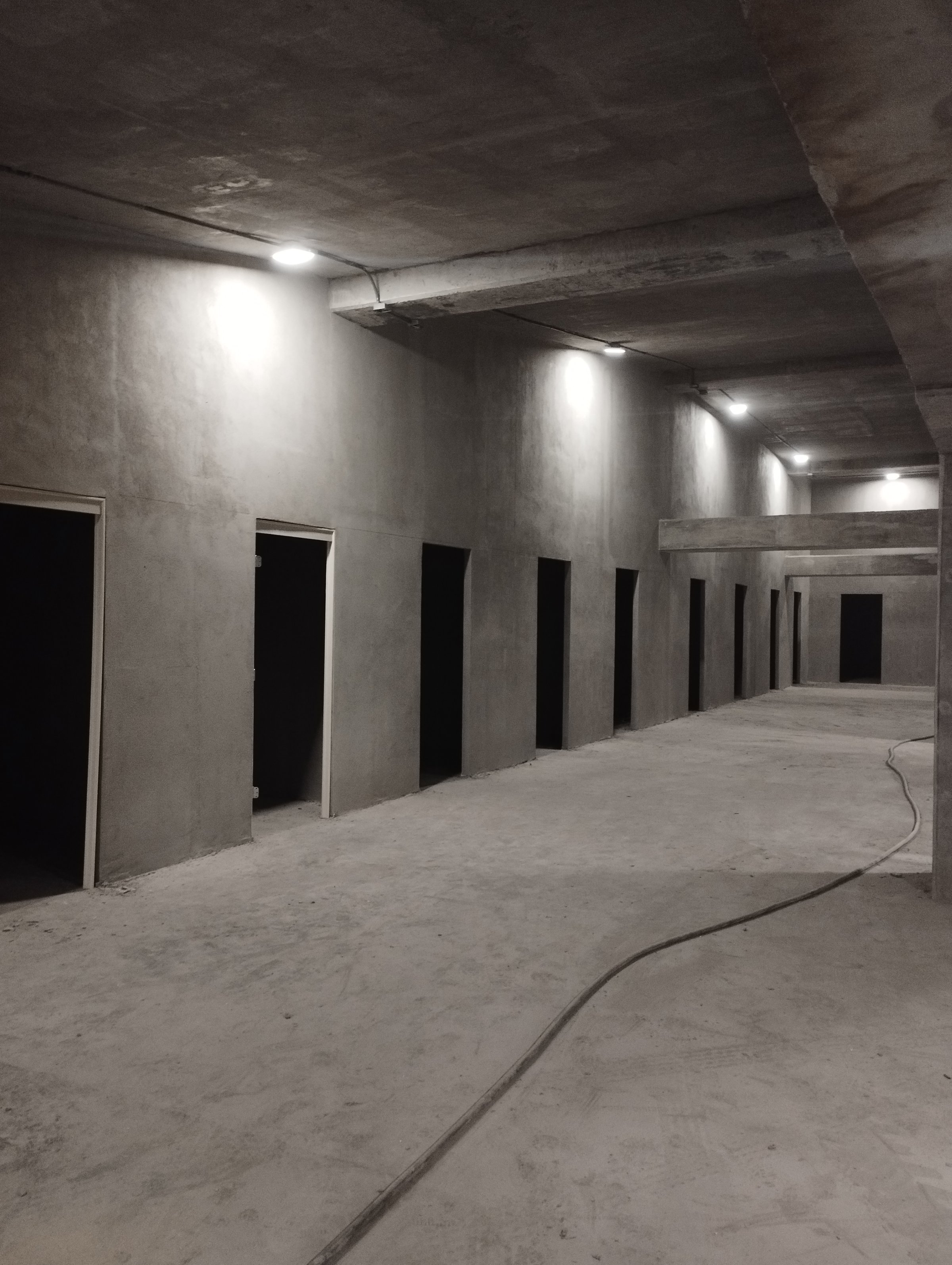Dimly lit, unfinished concrete corridor with open doorways on one side and a cable on the floor.