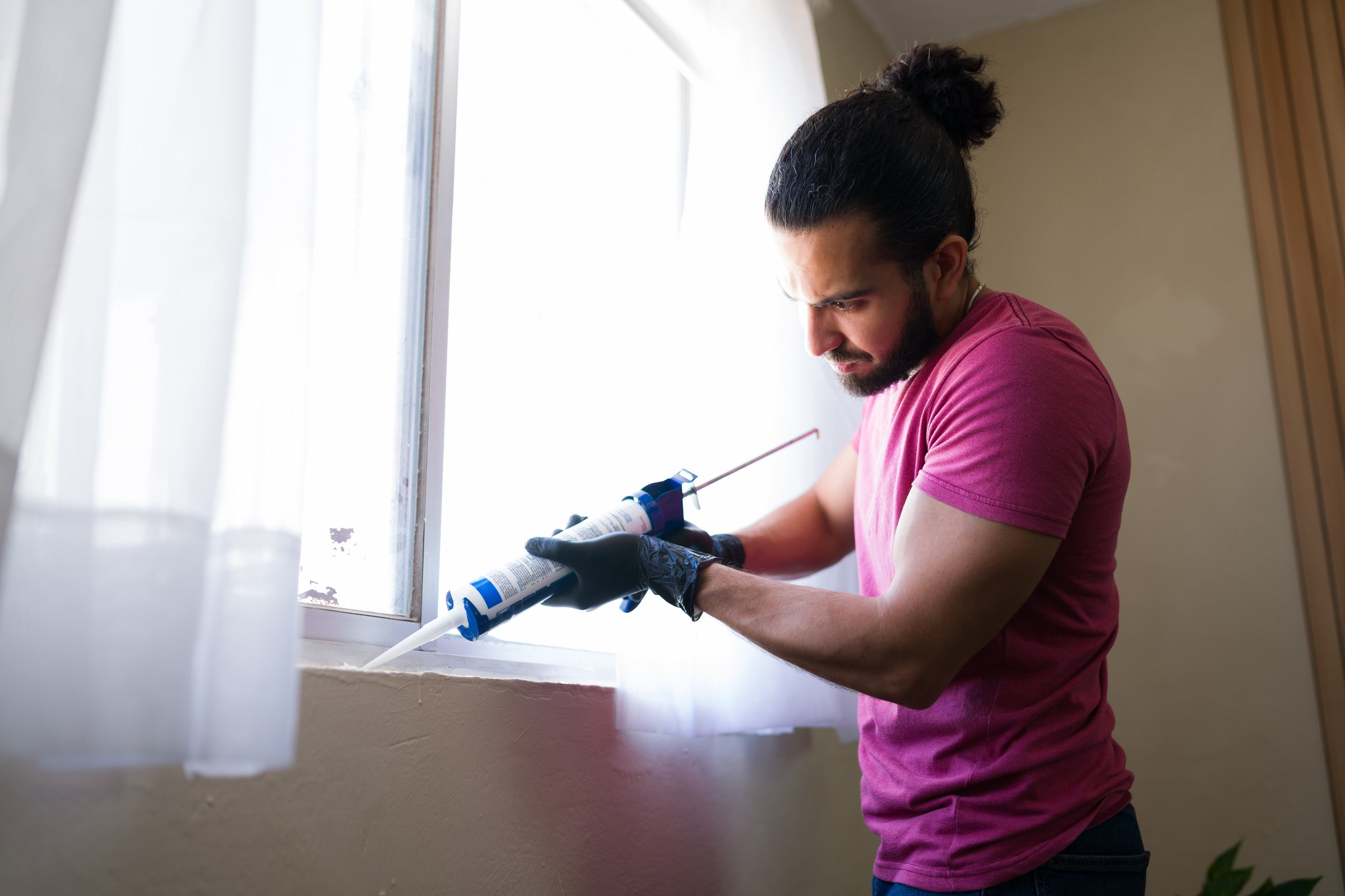 Handyman wearing gloves carefully applying silicone sealant to window frame, performing home repairs and improvements