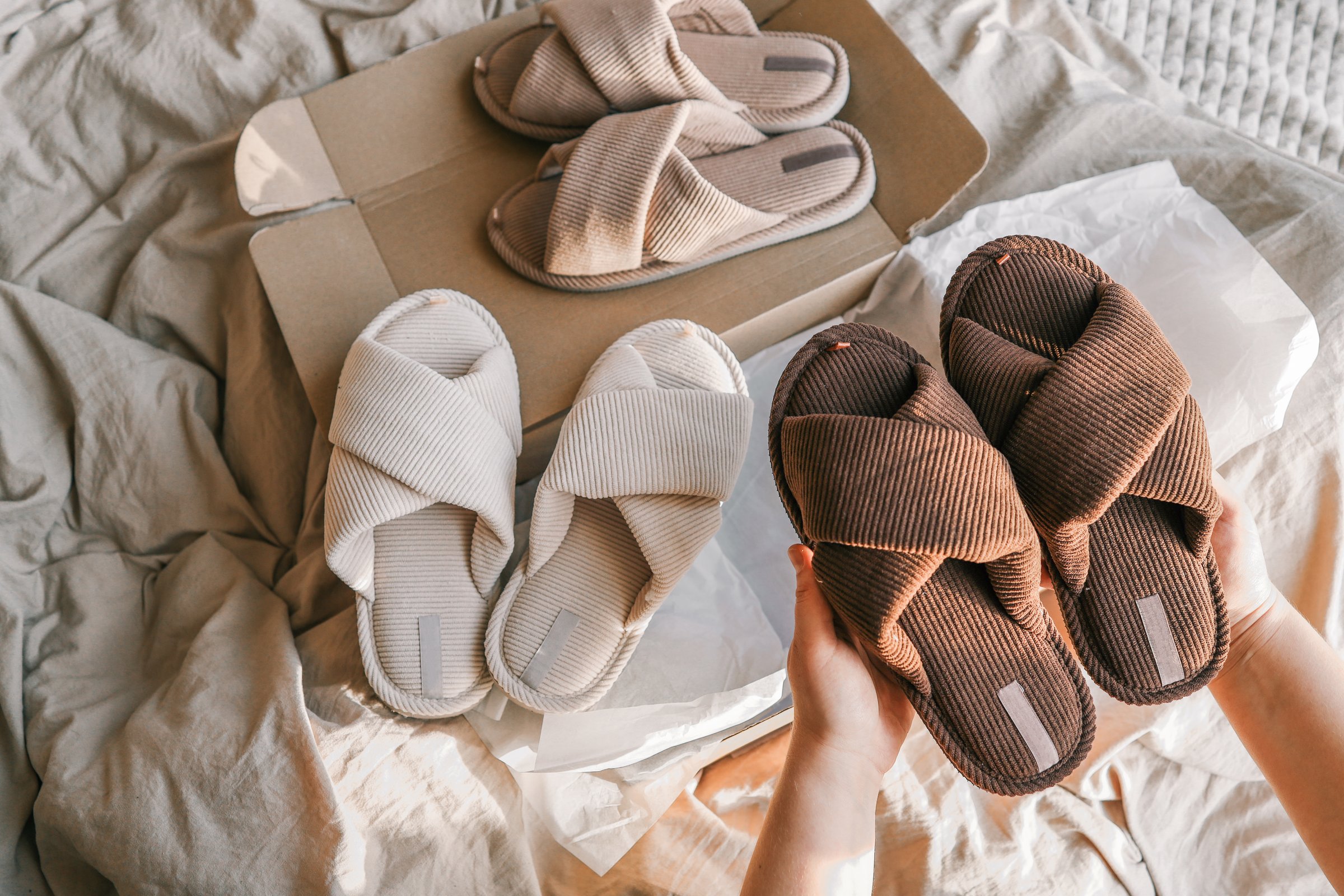 A collection of cozy corduroy slippers in earthy tones arranged on soft fabric with a cardboard box, bathed in warm natural sunlight, creating a relaxing and minimalist aesthetic.