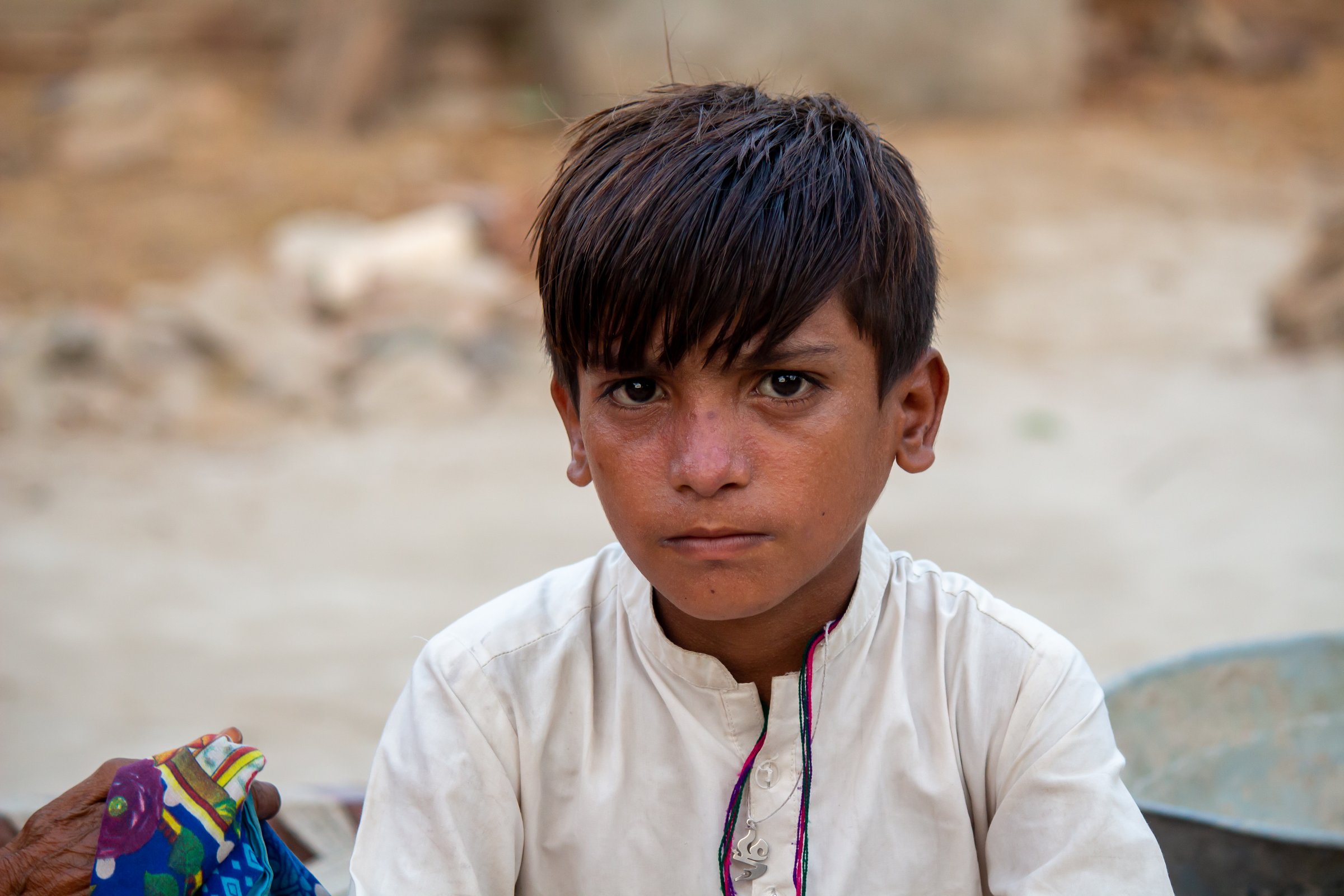 A young boy sits in a refugee camp, wearing a white shirt and looking thoughtful.