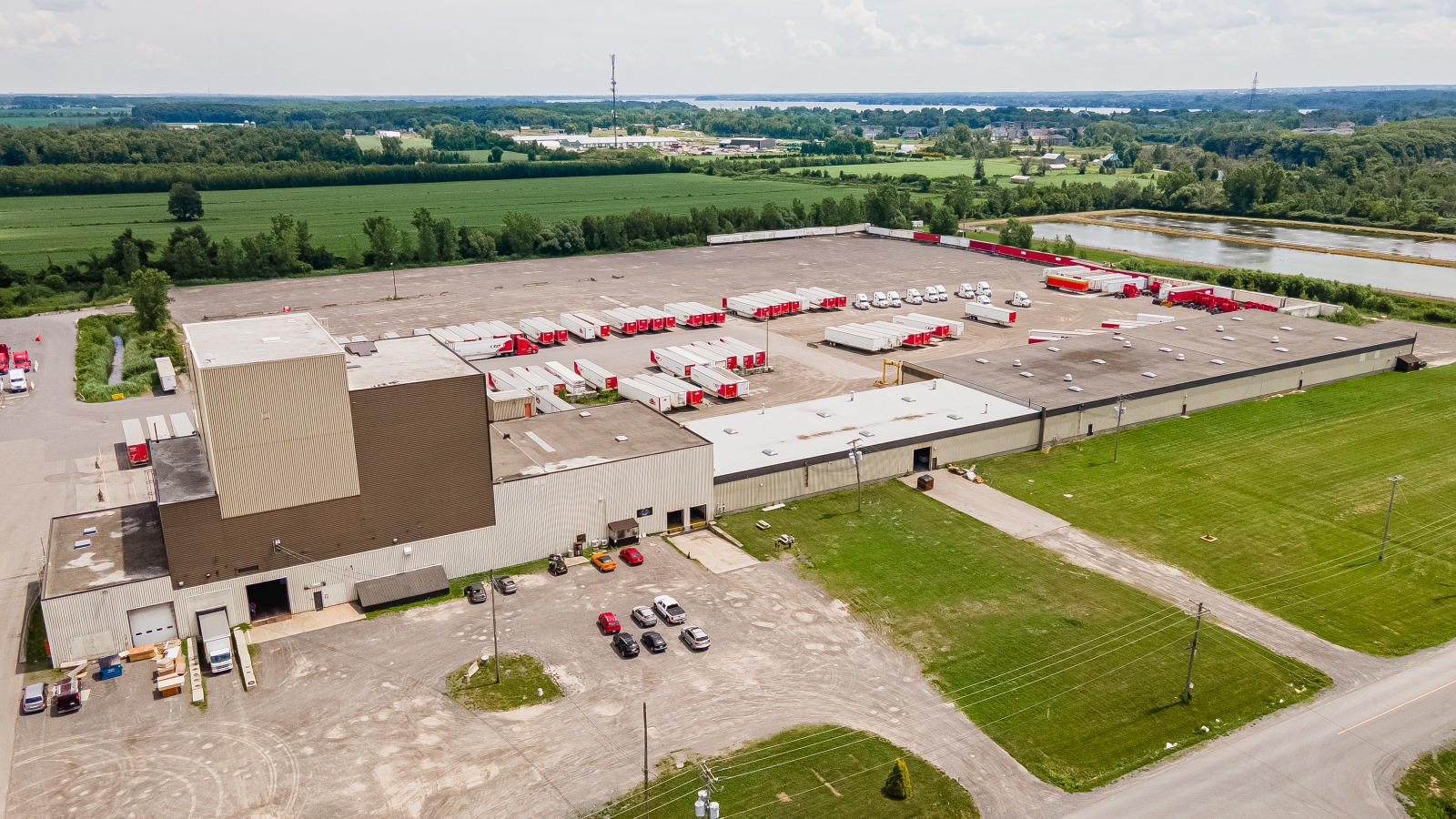 Aerial view of a large industrial warehouse with red trucks, surrounded by greenery and open land.