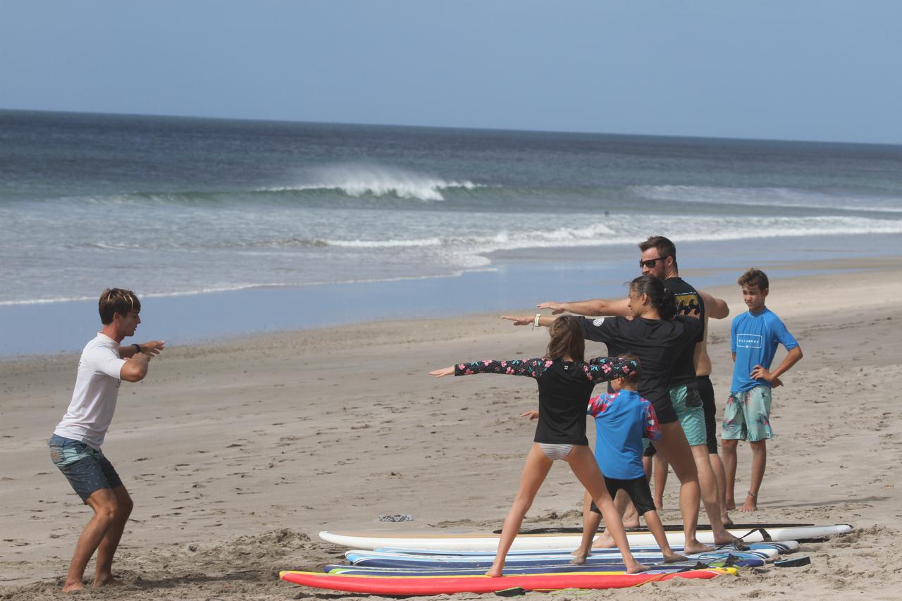 Surf instructor teaching a group of people how to balance on surfboards at a sandy beach with waves in the background.