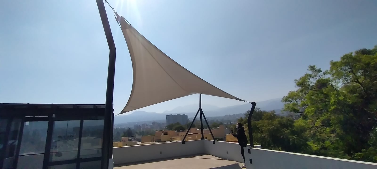 Outdoor rooftop area with a triangular shade sail canopy; a person adjusts its corner on a sunny day.