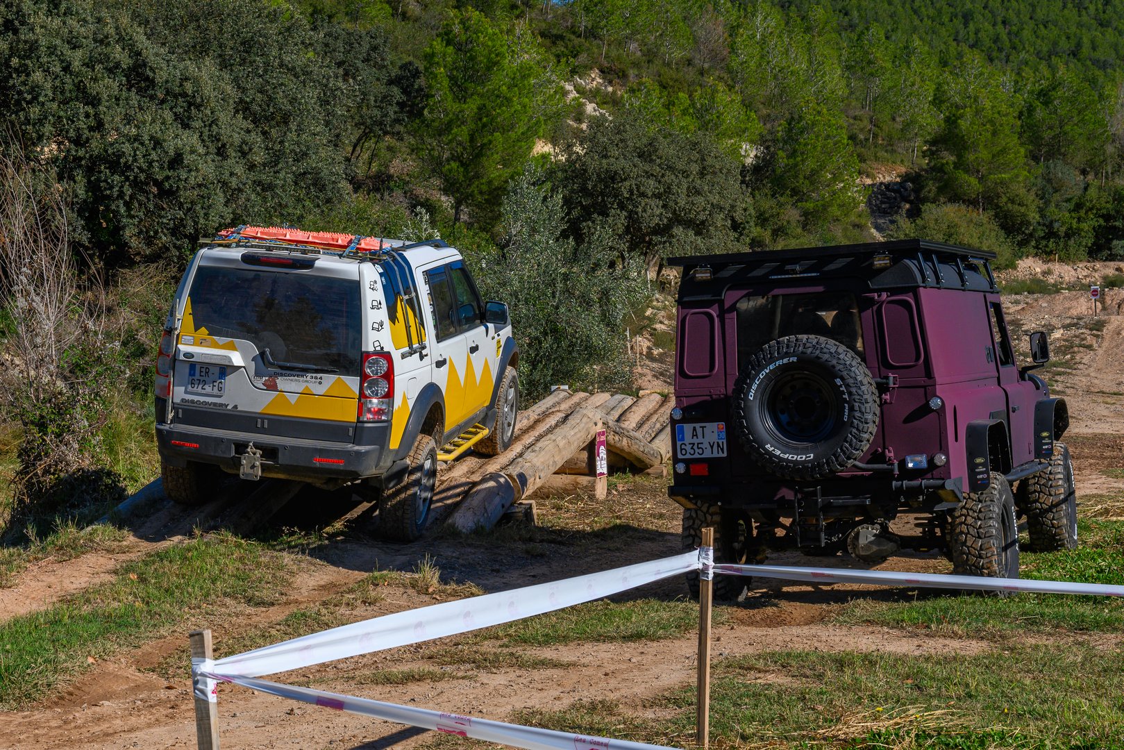 Land Party 2024, Les Comes, Spain 01/10/2024: Two Land Rovers, a white and yellow Discovery 3 with recovery boards on the roof, crossing a wooden bridge challenge. To its right, a maroon or dark wine-colored Land Rover Defender 110 with a black roof. Both vehicles have roof racks, and the second one has a spare tire mounted on the rear door. They are on an off-road test circuit.