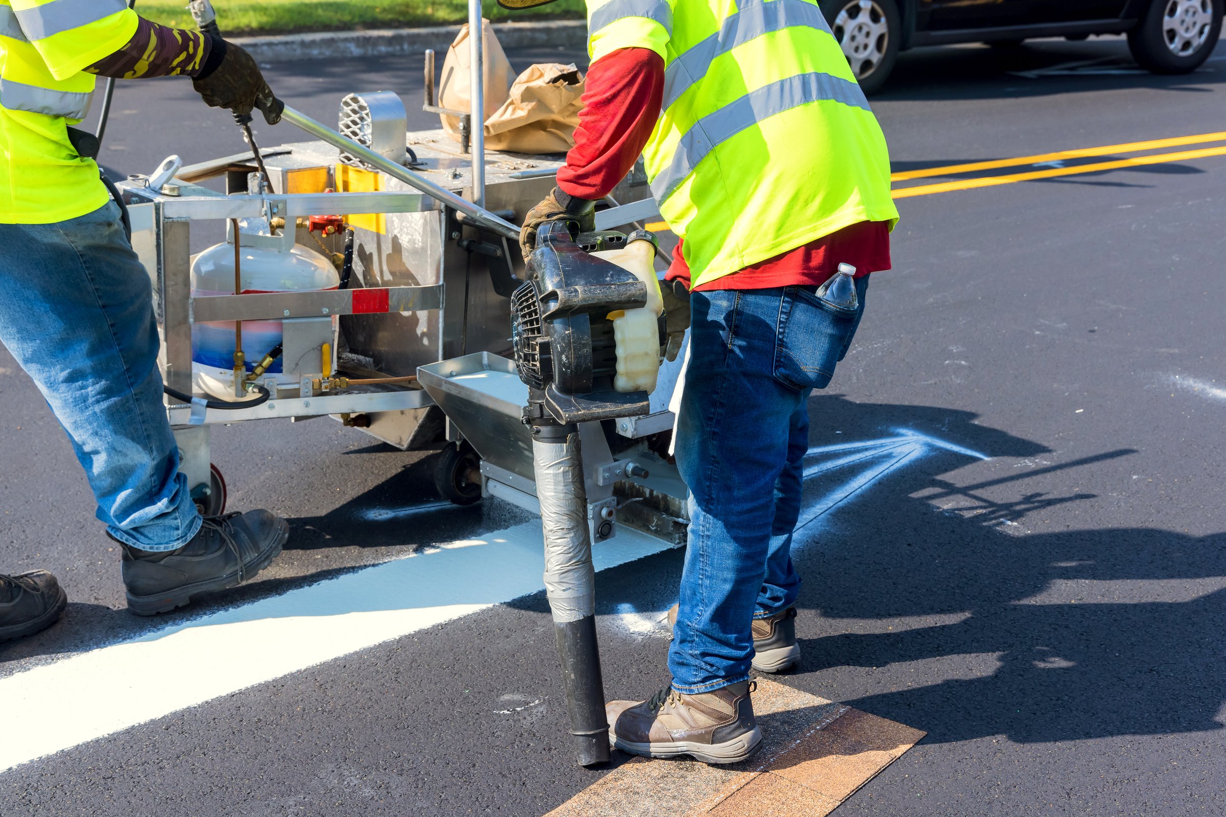 Workers are painting white road lines on an asphalt surface while traffic passes by under clear skies.