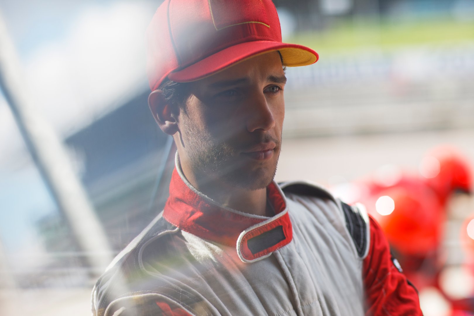 Race car driver in a red and white suit and cap stands focused at a racetrack, with blurred racing helmets in the background.