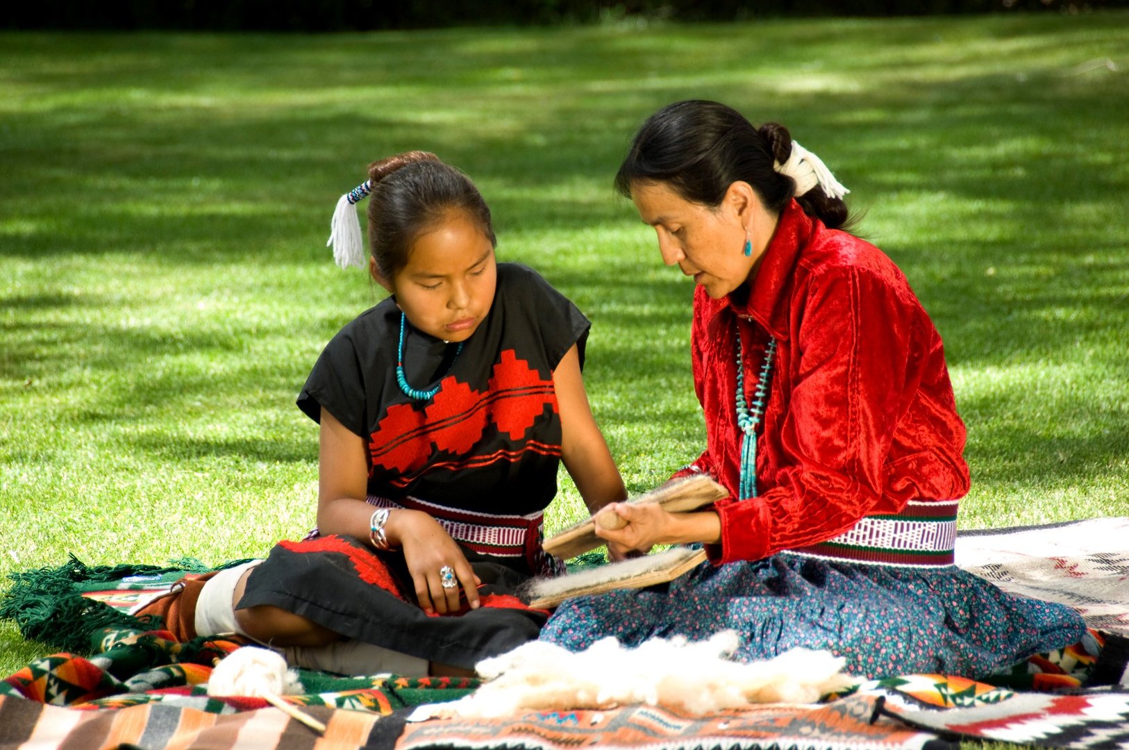 Two people in traditional attire sit on the grass, engaged in craftwork on a patterned blanket.