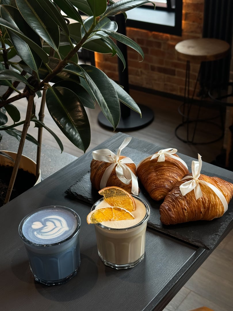 A tray on a table in a coffee shop with three fresh croissants wrapped with a beige ribbon, a glass of blue matcha and a glass of latte with orange slices.