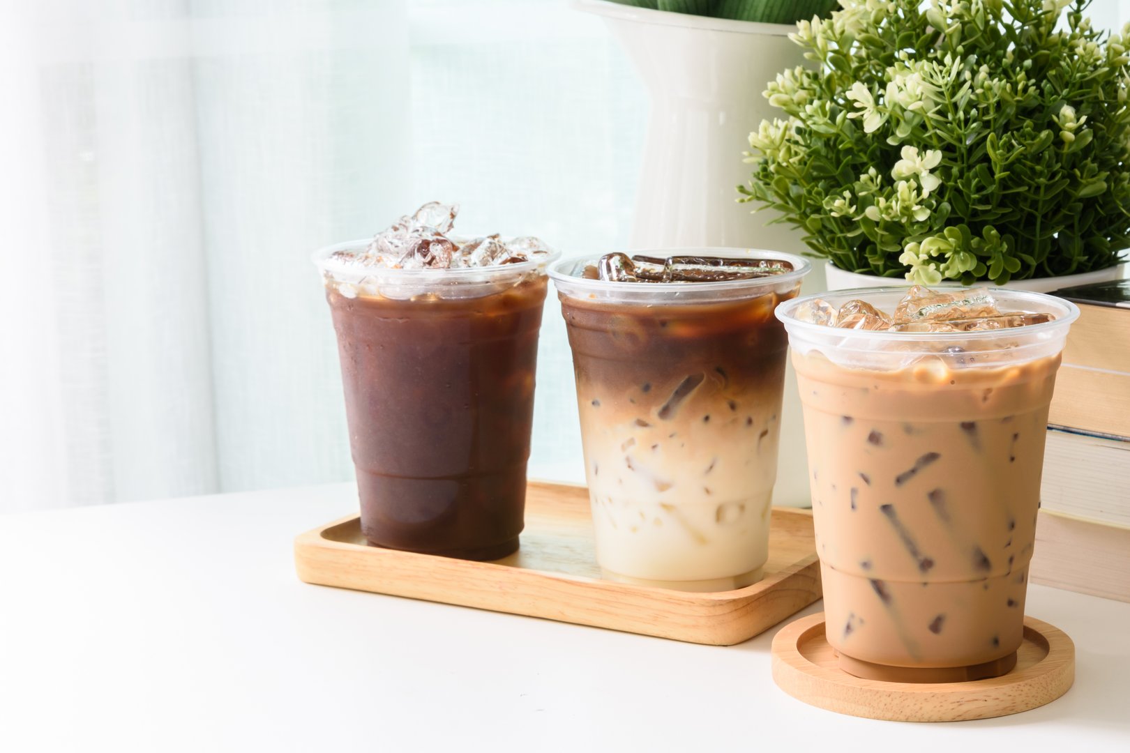 Ice americano, Ice espresso, Ice latte coffee, and a wooden tray on a white table indoors with white curtain background decorated with flowers.