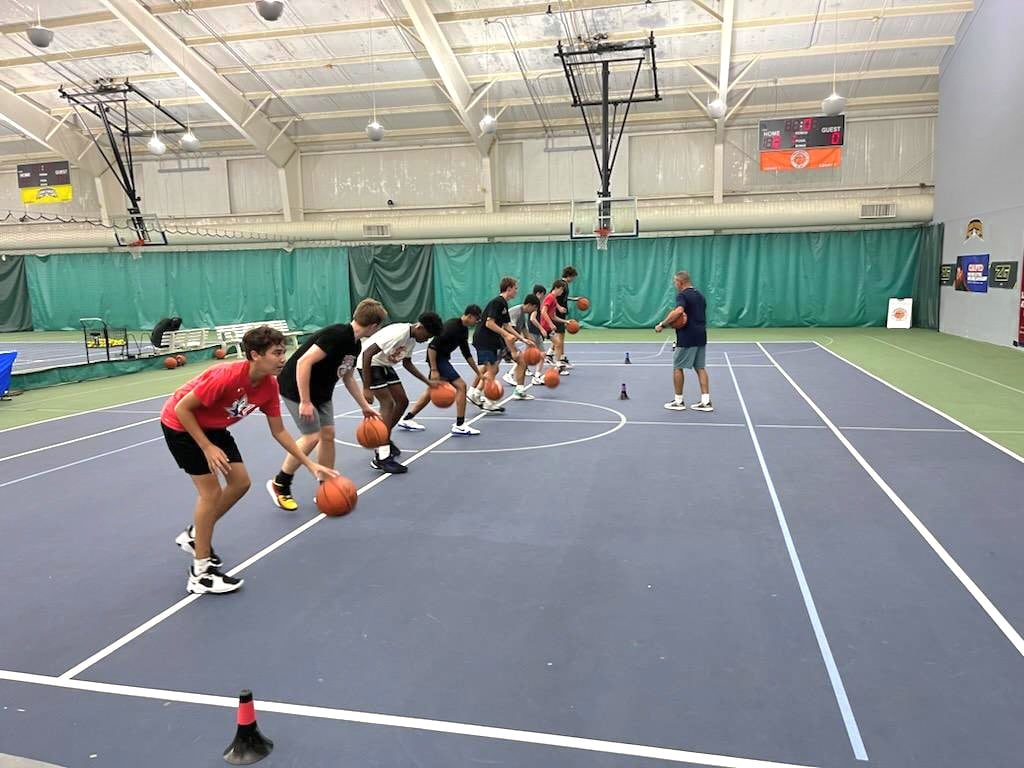 A group of boys in athletic attire practicing dribbling skills in a gymnasium under the guidance of a coach.
