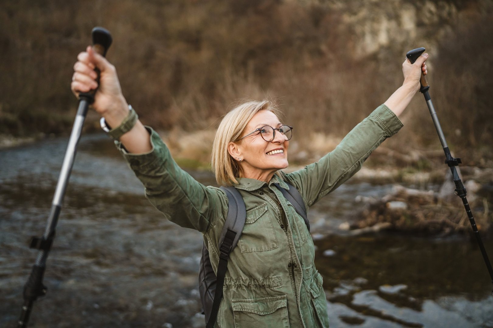 mature woman hiker backpacker explore forest and raise hands with hiking sticks across the river
