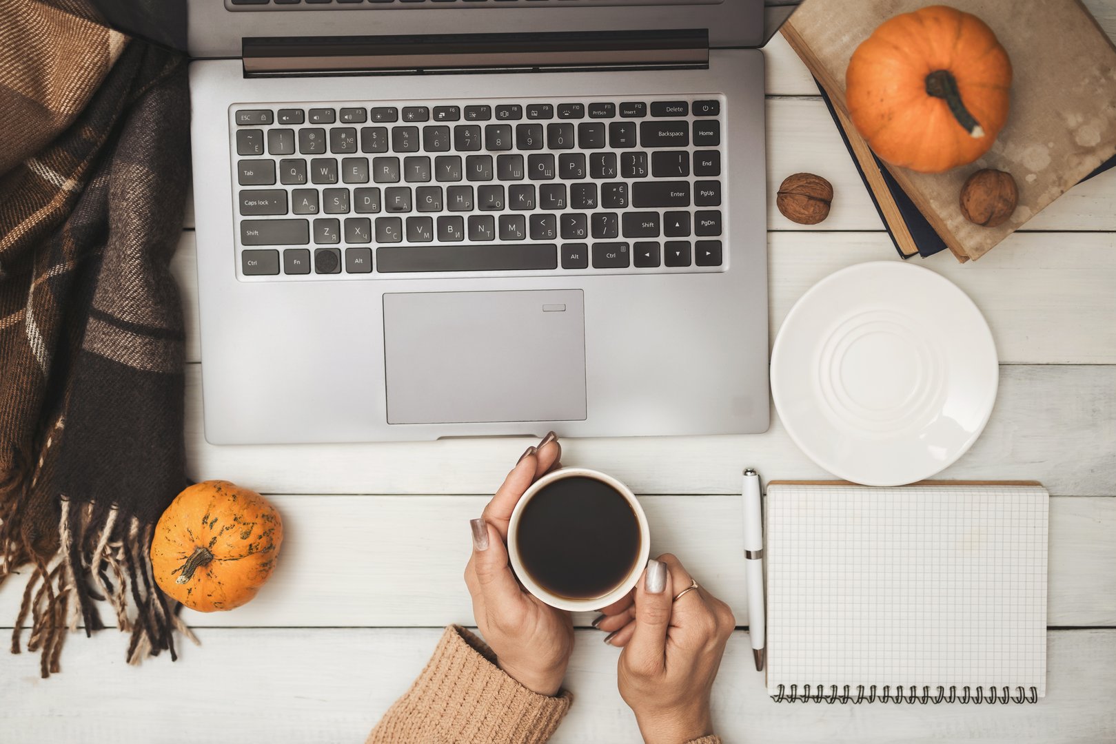 Female hands in sweater holding coffee cup and using laptop on white wooden background with pumpkins and notebook, cozy autumn workspace scene, top view.