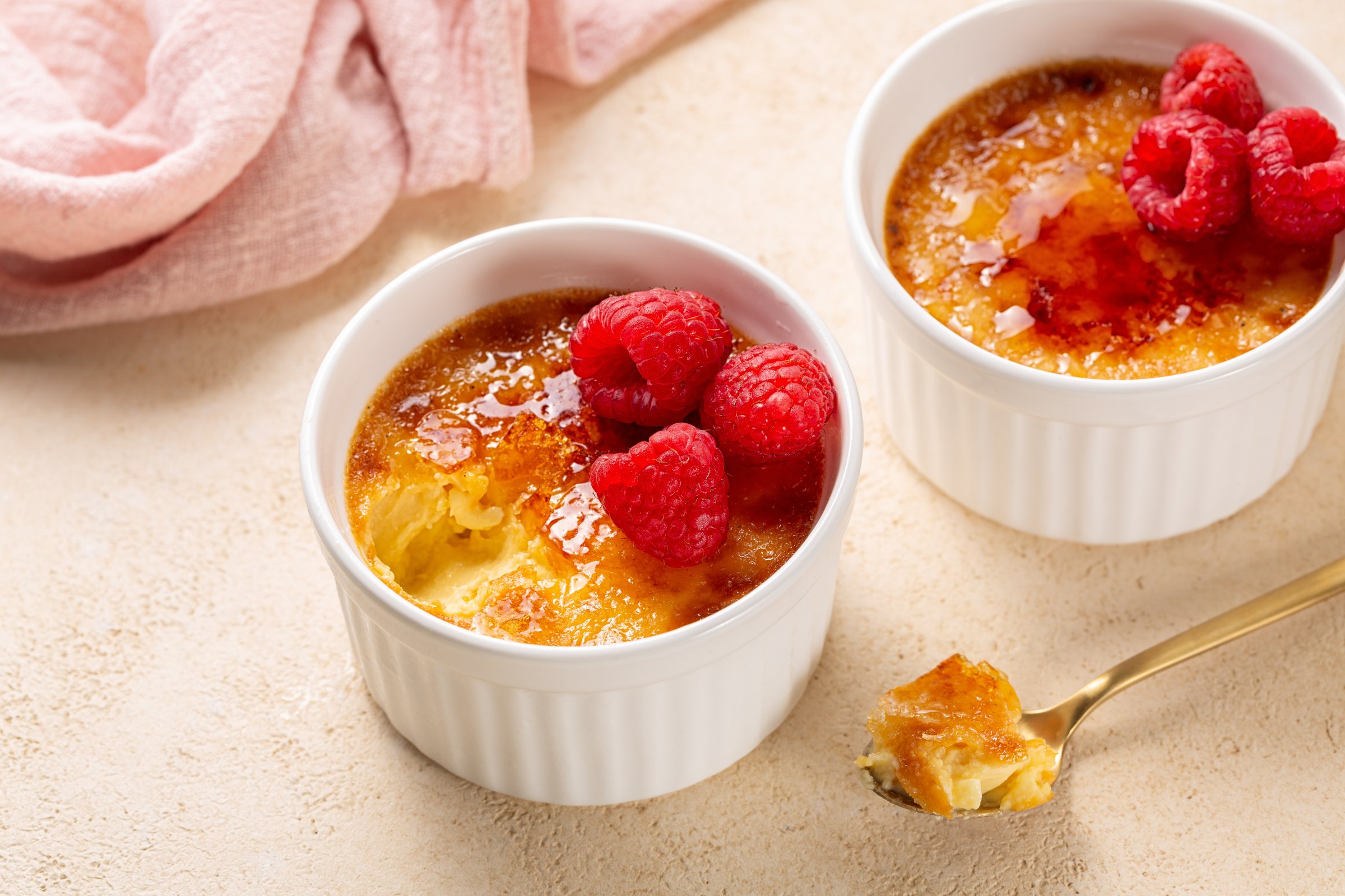 Eating Creme brulee, or burnt cream,  a dessert consisting of a rich custard base topped with a layer of hardened caramelized sugar, fresh raspberries. Two portion, Beige table surface.