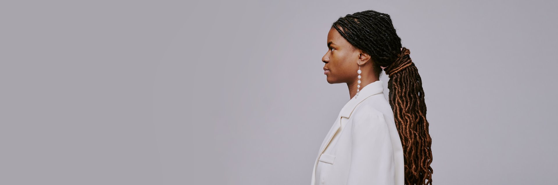 Woman with intricate braids wearing elegant white outfit in profile view against plain background showcasing refined fashion sense and composed demeanor