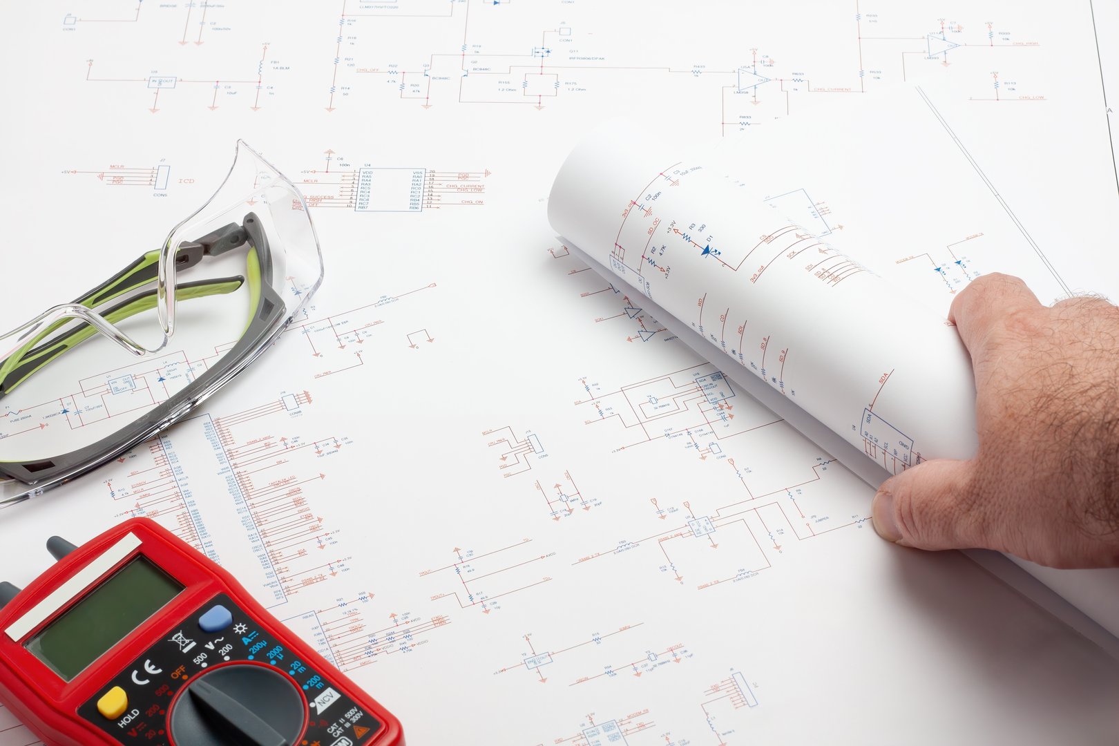 An electrical engineer's workspace featuring a hand holding a rolled blueprint, safety glasses, and a red multimeter on top of detailed electrical schematics. The image emphasizes the careful examination and measurement of electronic designs.