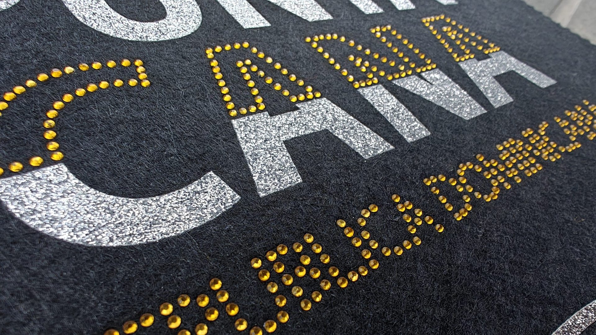 Close-up of textured letters on a dark surface, featuring glittery silver text and yellow rhinestone embellishments.