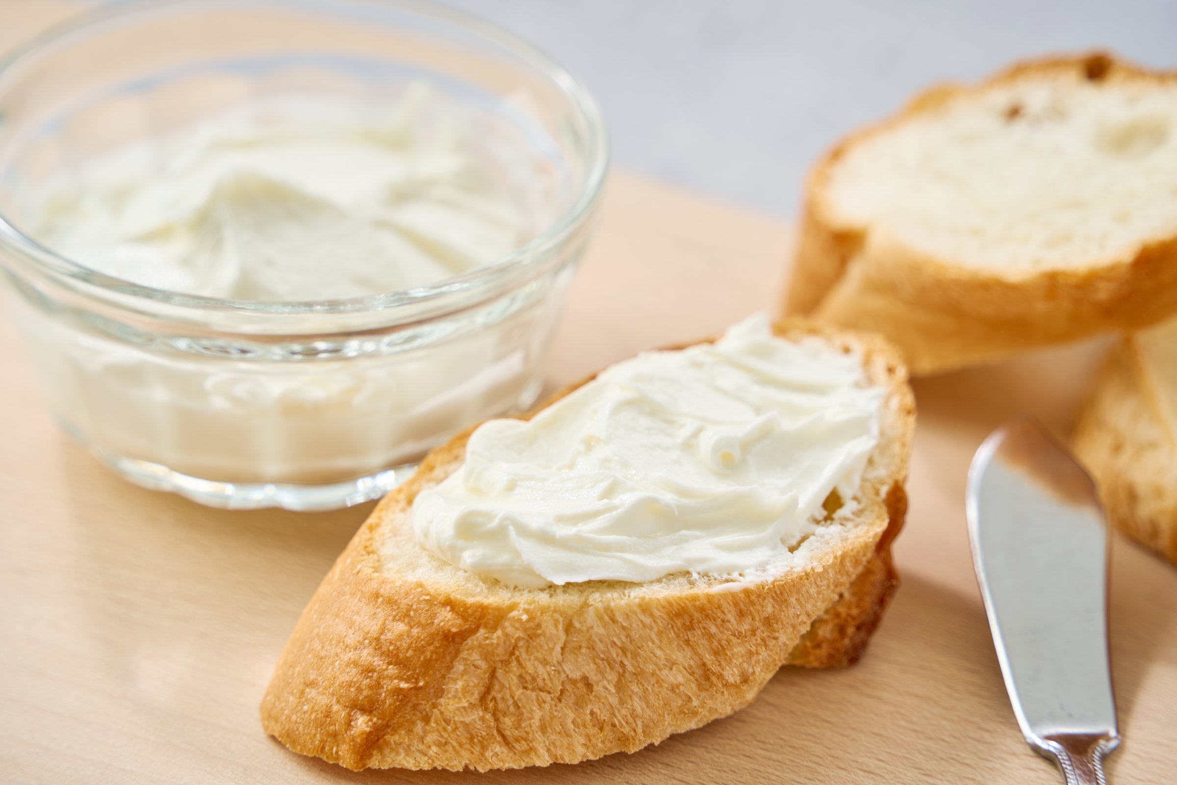 Fresh cream cheese spread on a slice of baguette, served with a glass bowl of cheese on a wooden board.