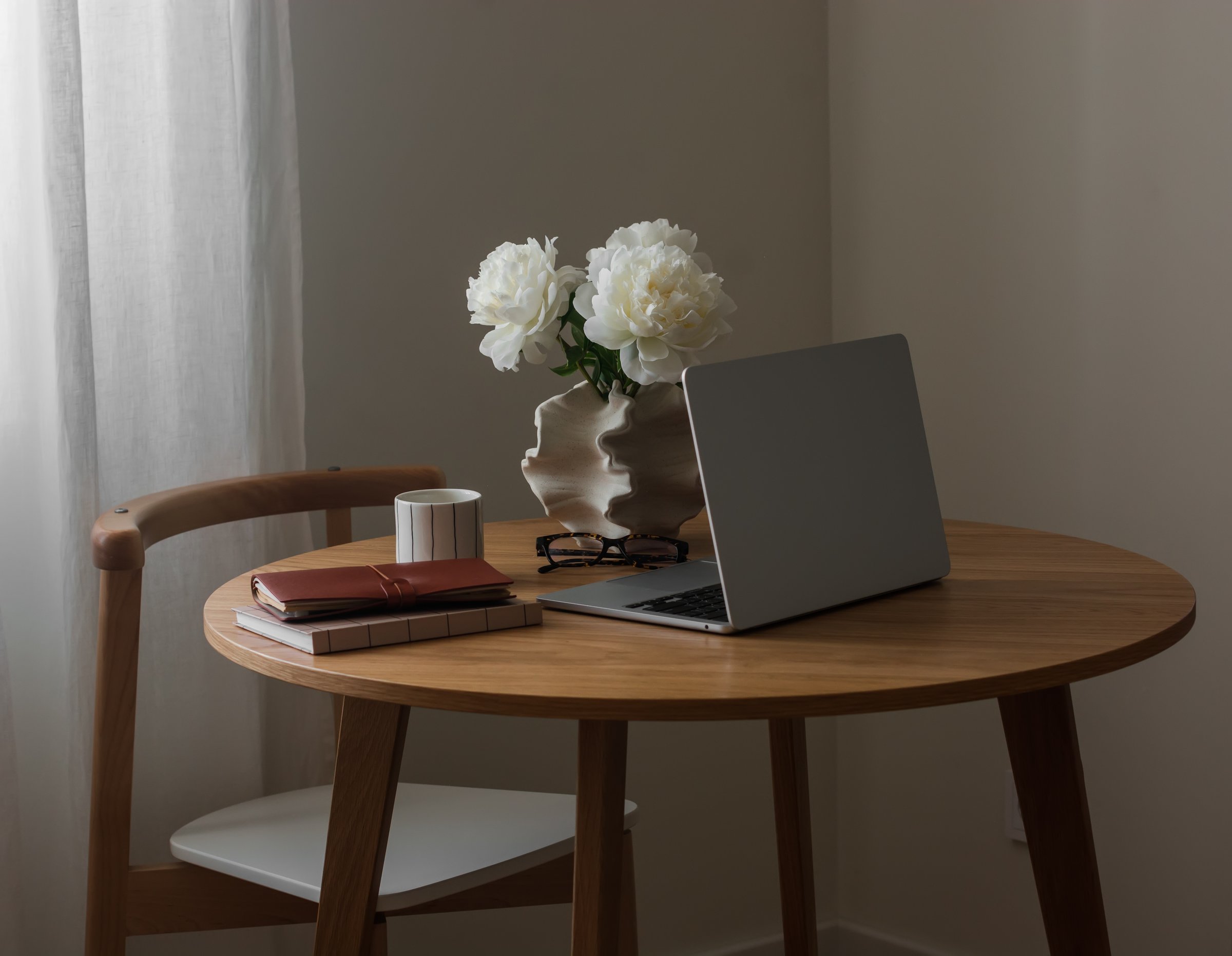 A laptop, a vase with peonies, a glass of coffee, books on a round wooden table in the living room