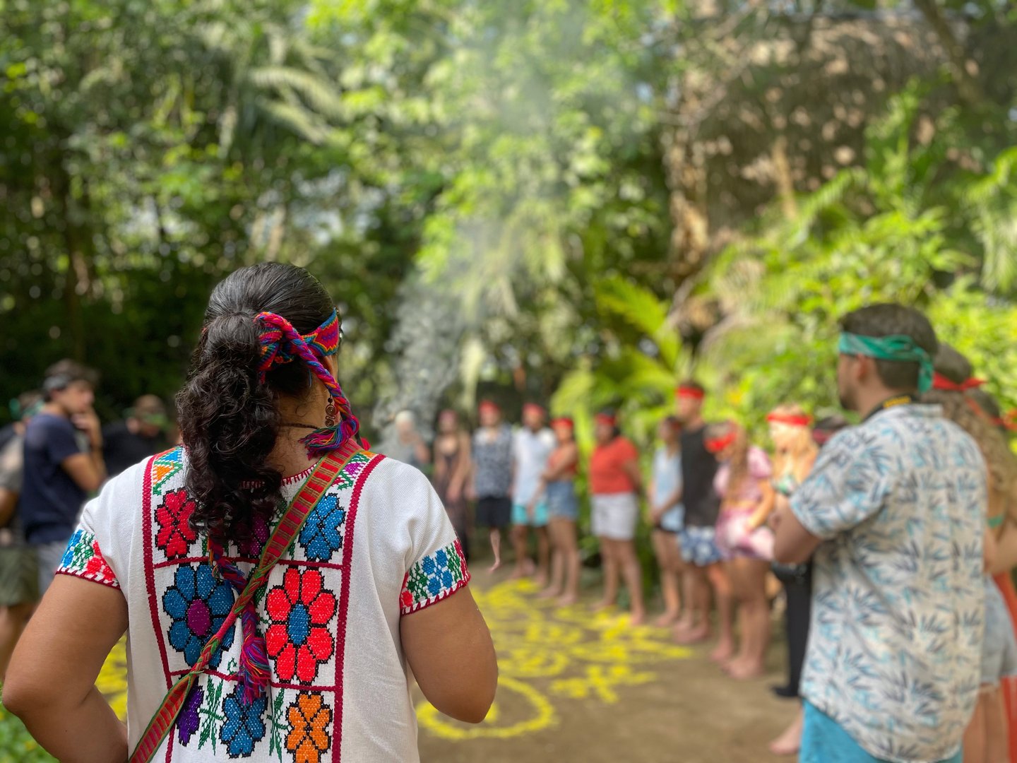 A woman in traditional attire leads a group ritual in a lush forest setting, with participants forming a circle.
