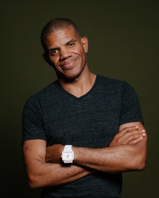 Smiling person with short hair wearing a black T-shirt and white watch, arms crossed against a dark green background.