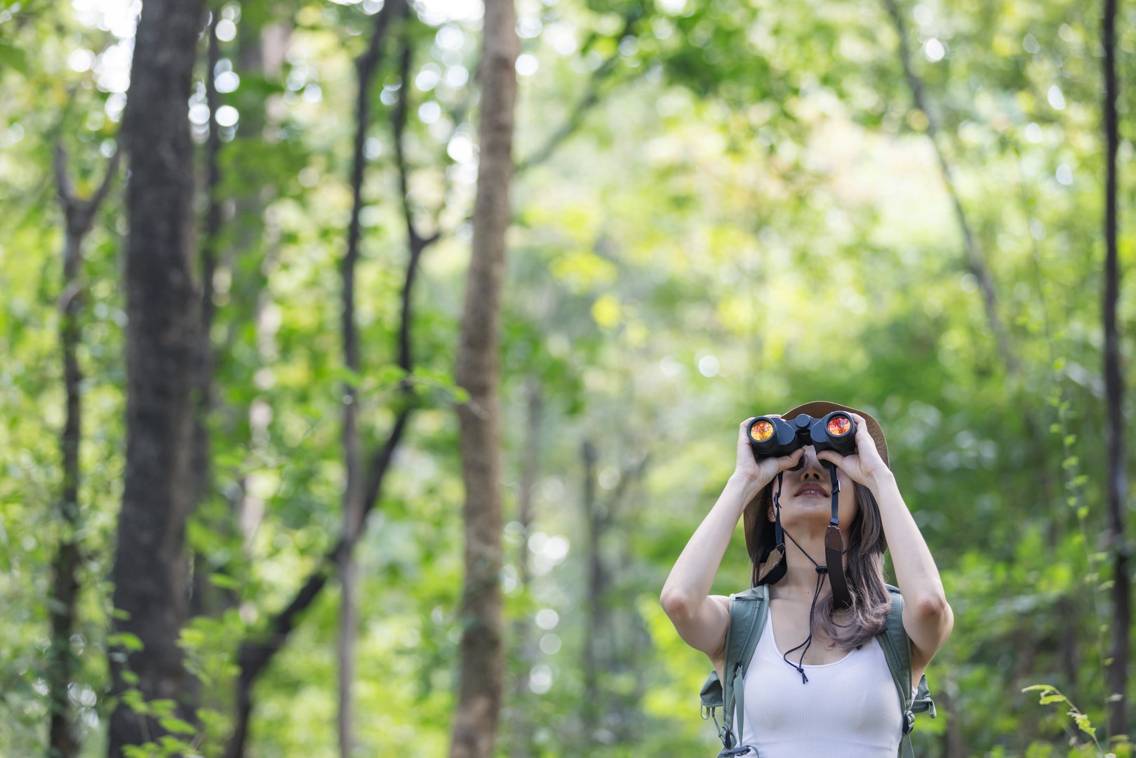 A young woman wearing a sun hat and light-colored outfit stands in a lush green forest, holding binoculars. She looks up with curiosity, as if observing birds or wildlife in the peaceful environment