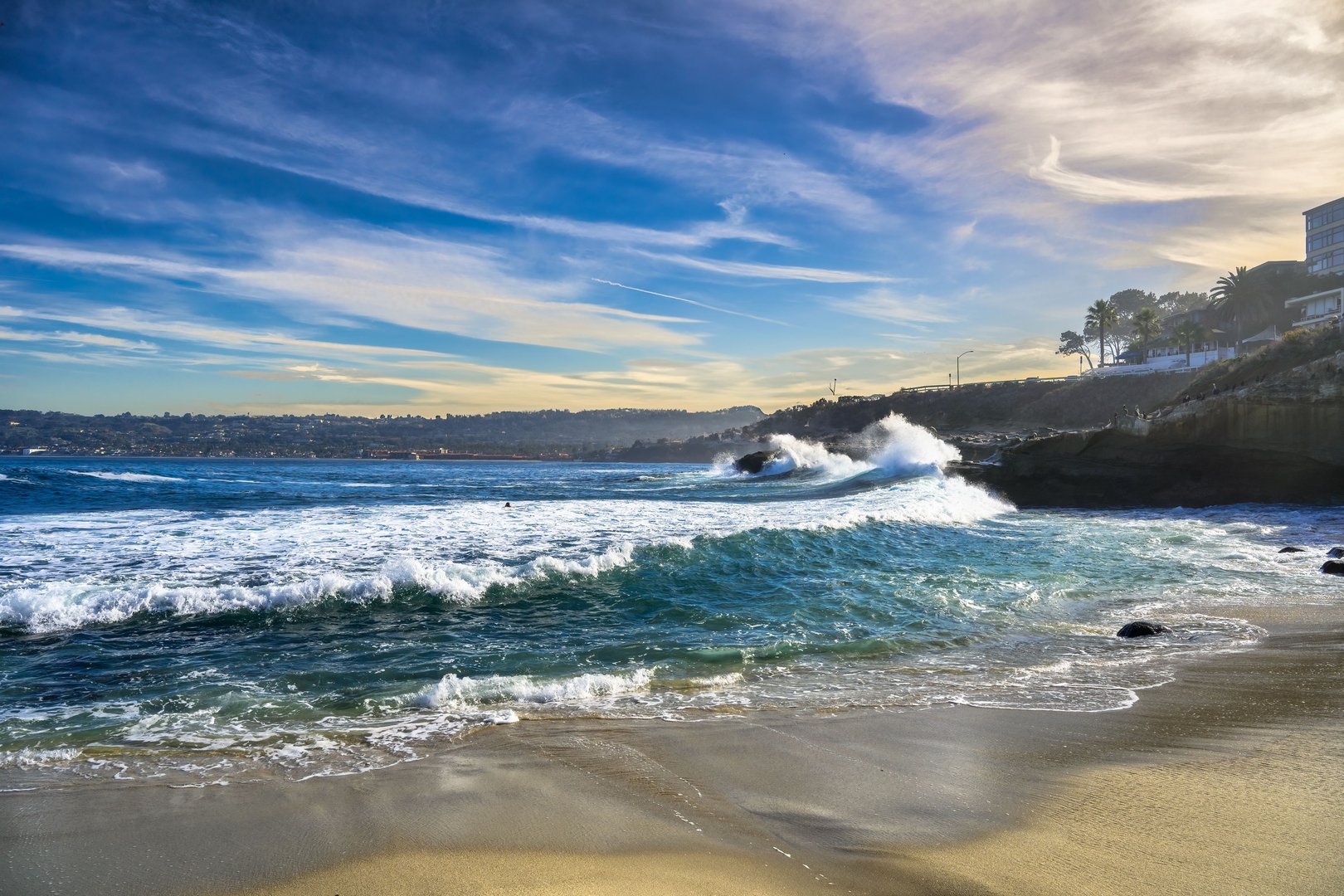 Waves crash against the rocks at La Jolla Cove, with water spraying and a light blue sky in California.