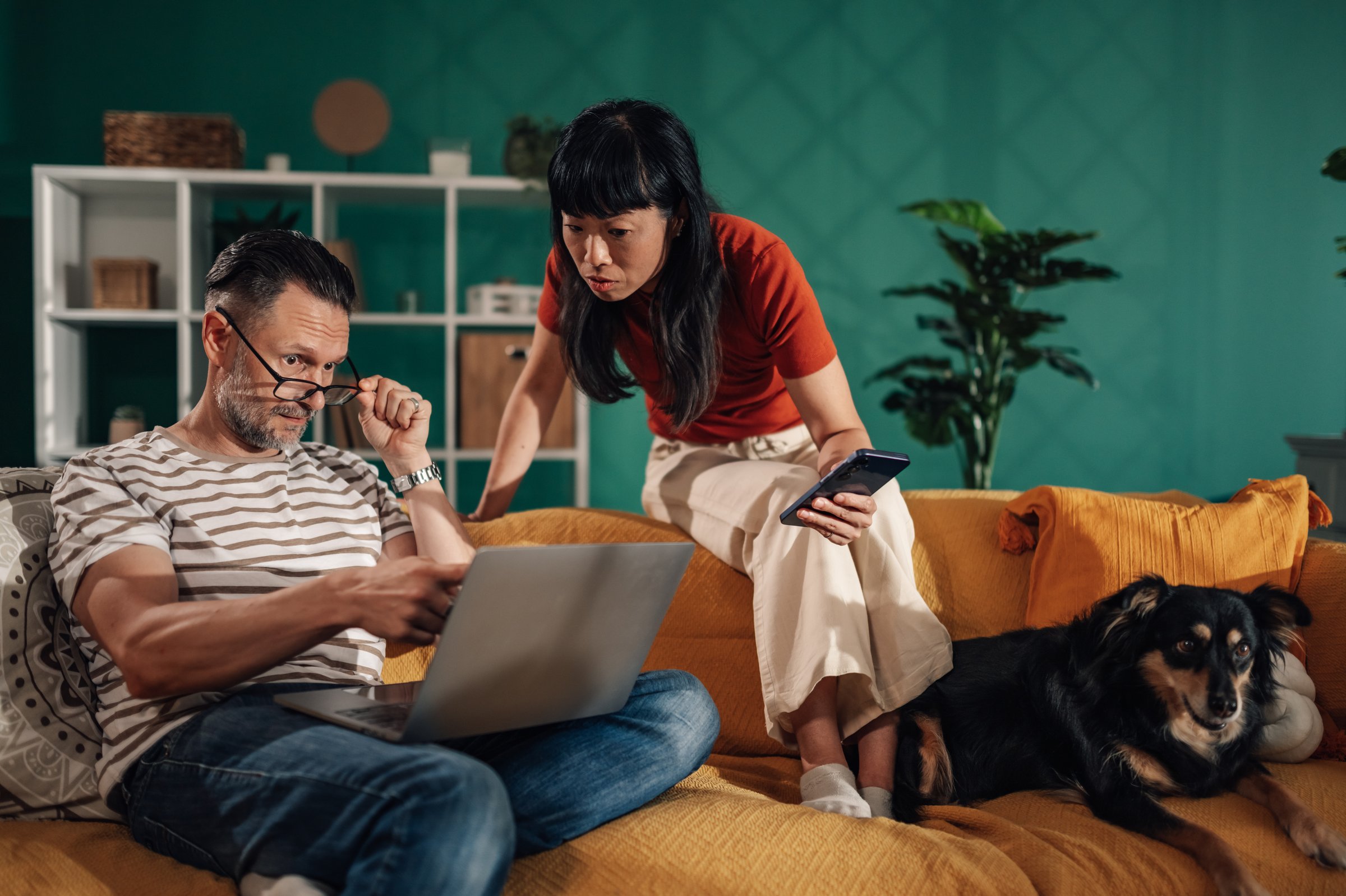 Couple sitting on sofa with their dog, browsing internet on laptop and smartphone, enjoying leisure time at home