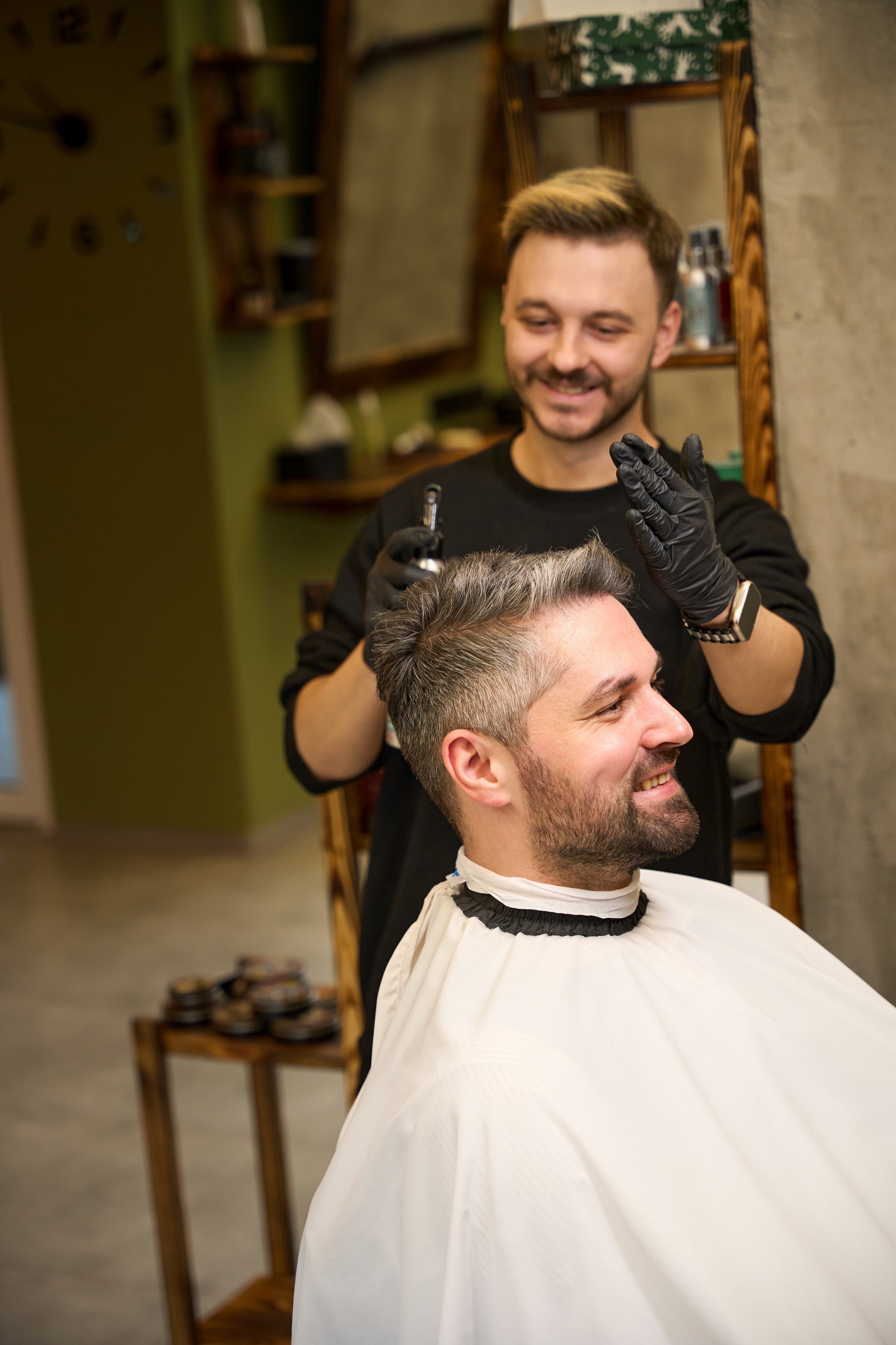 Positive bearded barber spraying fixing liquid into the haircut of handsome gray-haired man, satisfied client sitting in barber chair and looking at his reflection with delight
