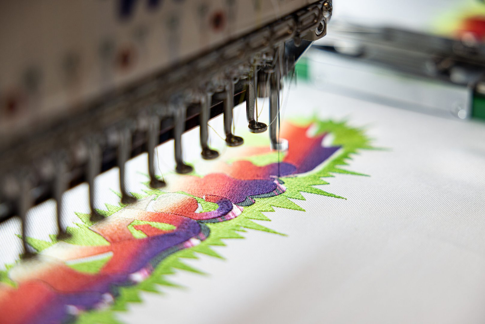Close-up of a machine embroidering a colorful, wavy pattern with green, purple, and red threads on fabric.