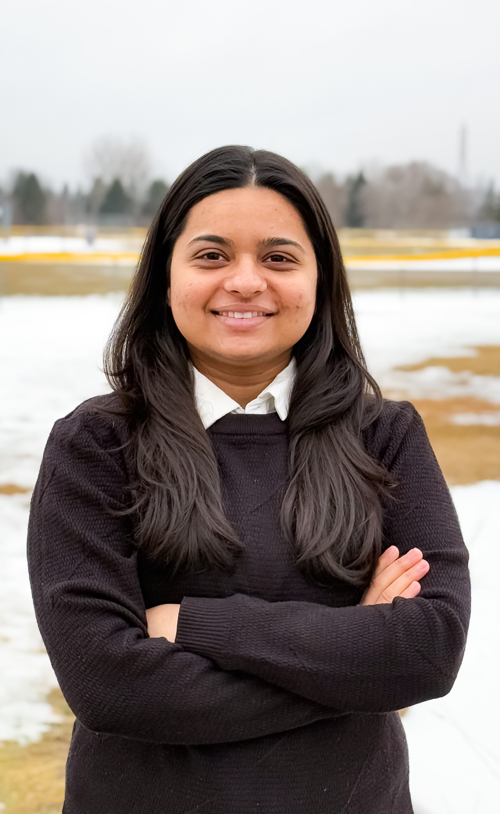 Person with long hair and a black sweater stands outdoors with arms crossed, snowy ground and trees in the background.