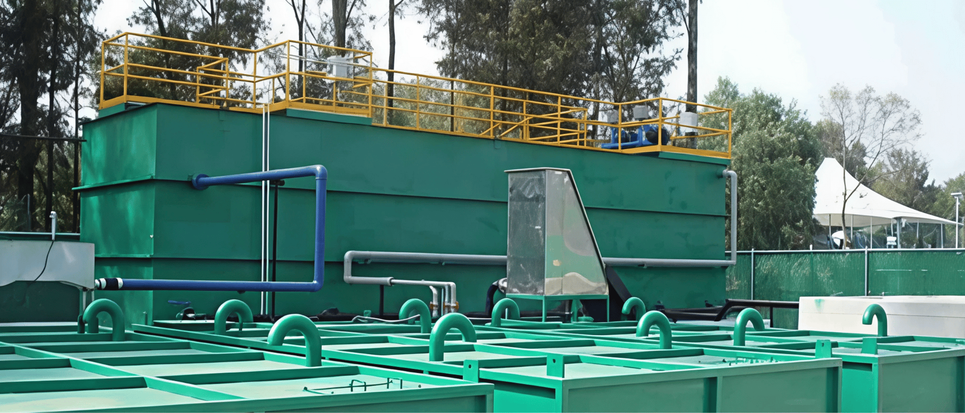 Green industrial tanks with pipes and railings, surrounded by trees and equipment in an outdoor setting.