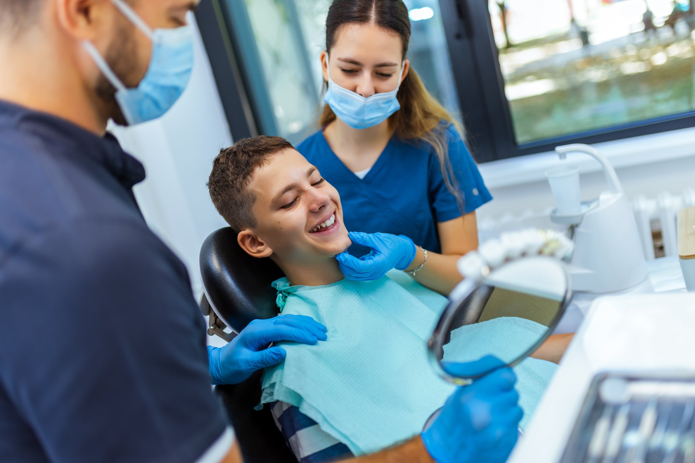 Little boy sits in dental chair, smiling happily as doctor and nurse hold a mirror to show his teeth, demonstrating joyful pediatric dental care in a modern clinic.