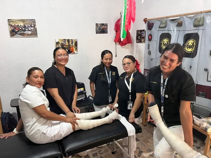 Five women in a room, two sitting on a medical table with leg bandages, others standing around smiling, wearing uniforms.