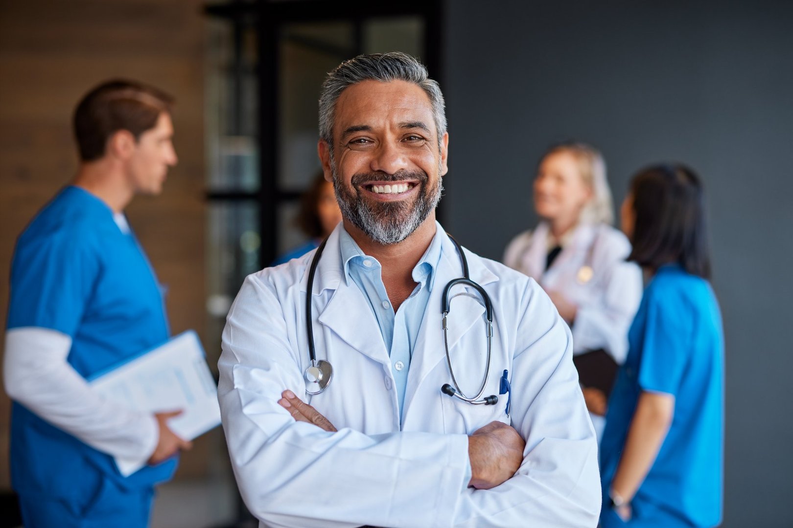 Confident indian doctor smiling with arms crossed while looking at camera with team in background. Medical middle eastern physician looking at camera with collaborative medical staff in background. Portrait of indian doctor standing proudly with colleagues discussing in the background.