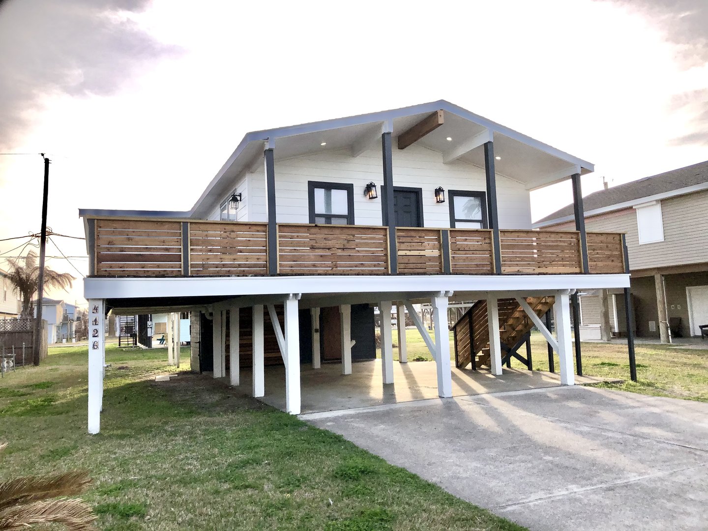 Elevated house with wooden balcony and stairs, on a grassy lot, with a concrete driveway under the structure.