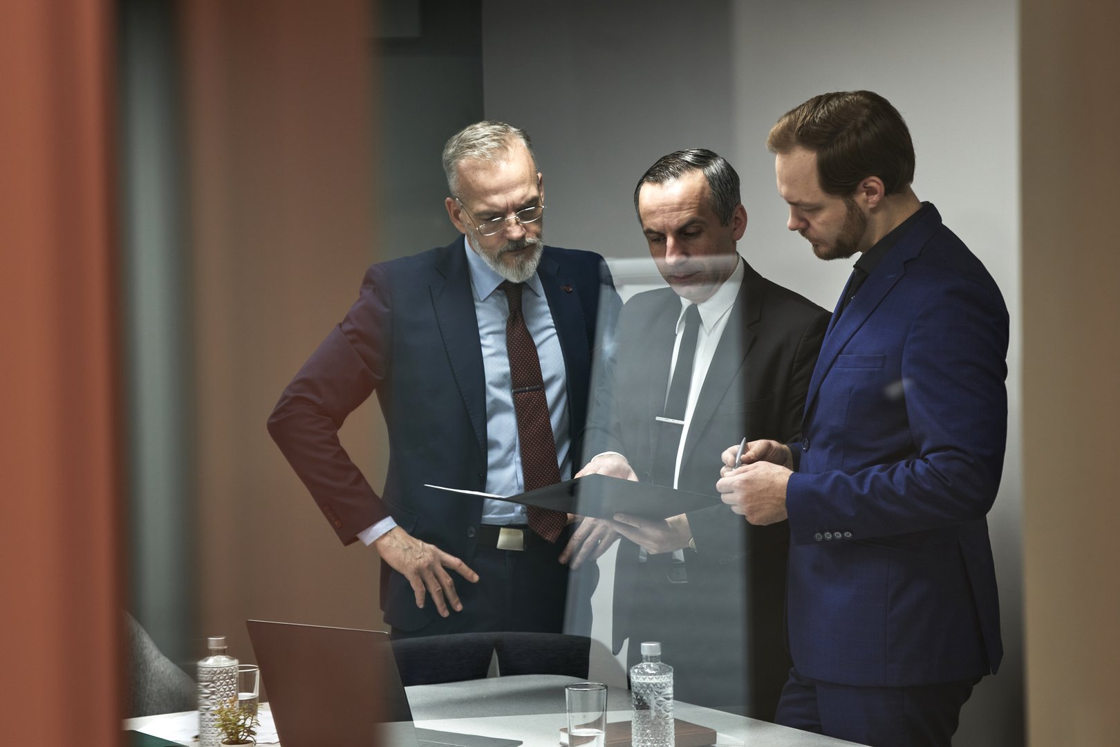 Three middle aged Caucasian men standing together discussing documents in modern office meeting room, two men listening while third man holding folder and gesturing with pen