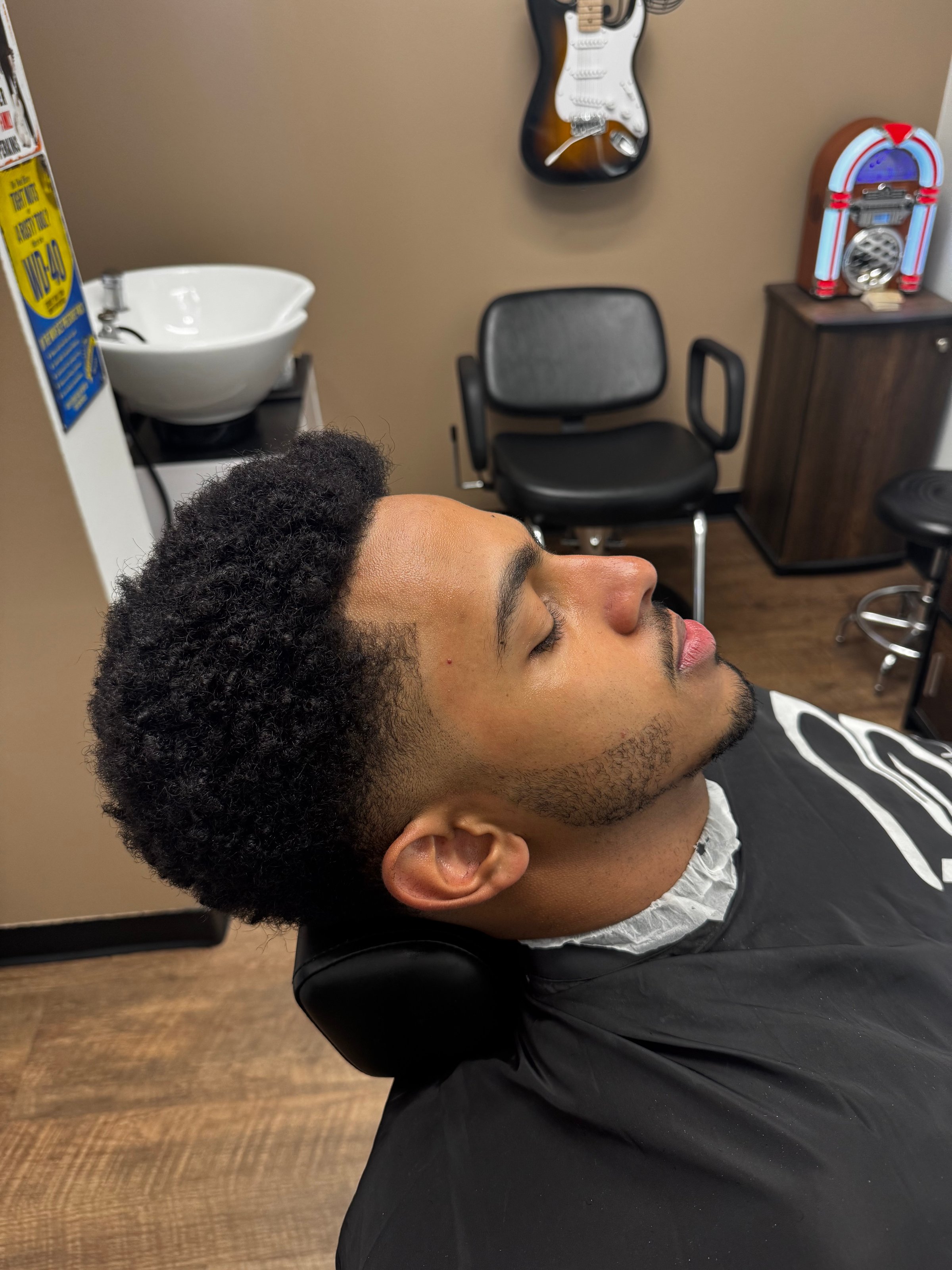 A man with a fresh fade haircut sits in a barber chair, with a guitar and jukebox in the background.