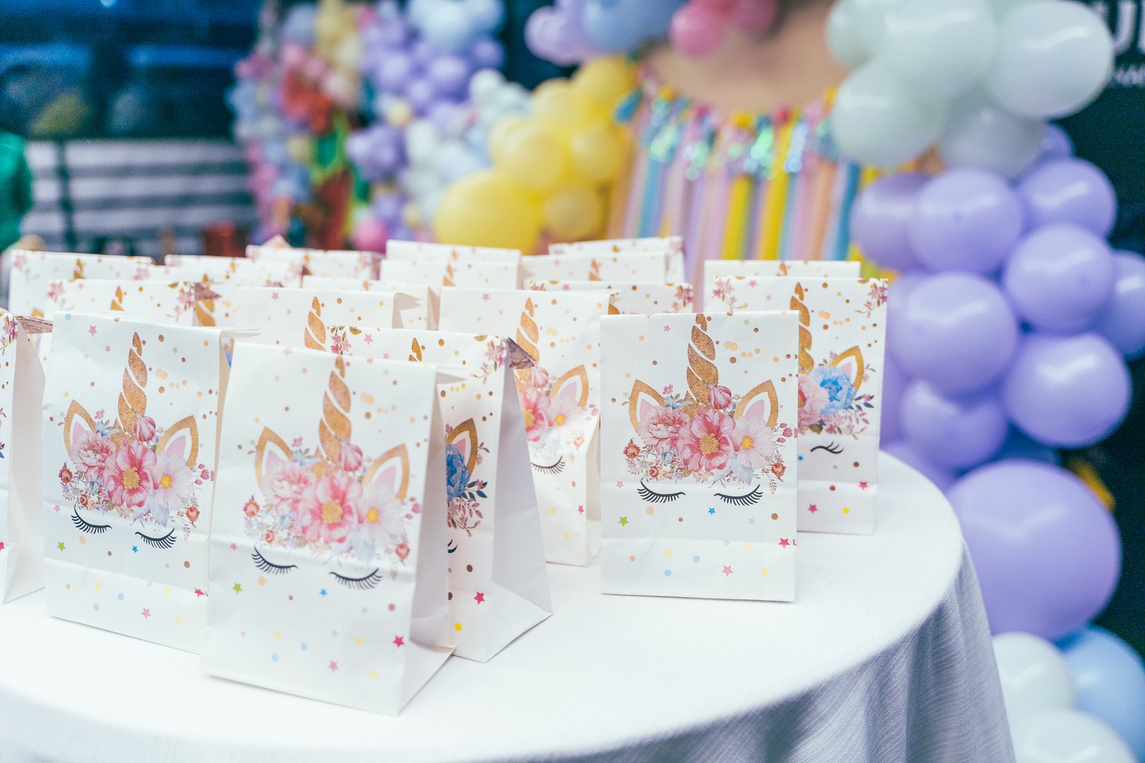 Unicorn-themed gift bags arranged on a round table, set up for a children's birthday party. Colorful balloon decorations in the background create a festive atmosphere