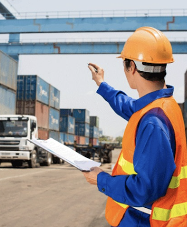 Worker in safety gear points at shipping containers, holding a clipboard at a busy freight yard.