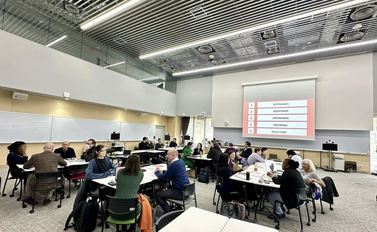 People engaging in group discussions around tables in a modern conference room with a presentation screen displaying text.