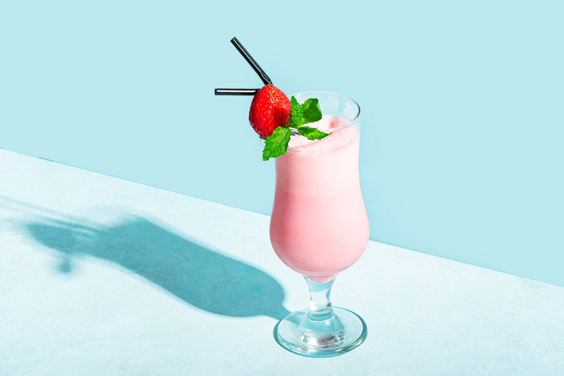 Glass of fresh strawberry milkshake, smoothie with mint and fresh strawberry on light blue background, hard shadow, copy space. Healthy food and drink concept. Berry protein shake.