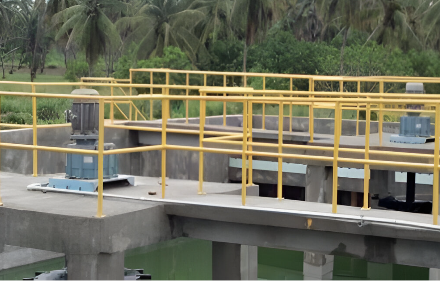 Industrial facility with yellow railings, machinery, and green water basin, surrounded by palm trees.