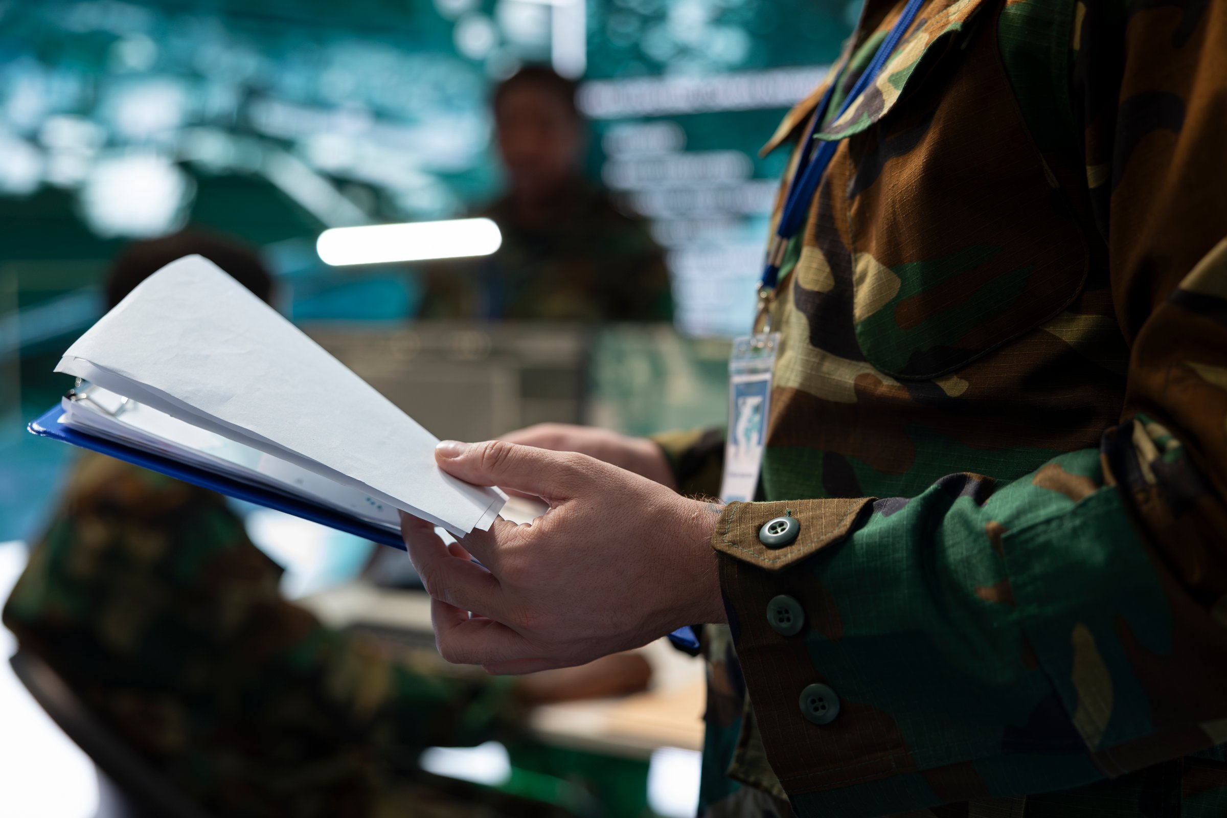 Military personnel in camouflage gear reviewing documents in a command center with computers and screens visible.