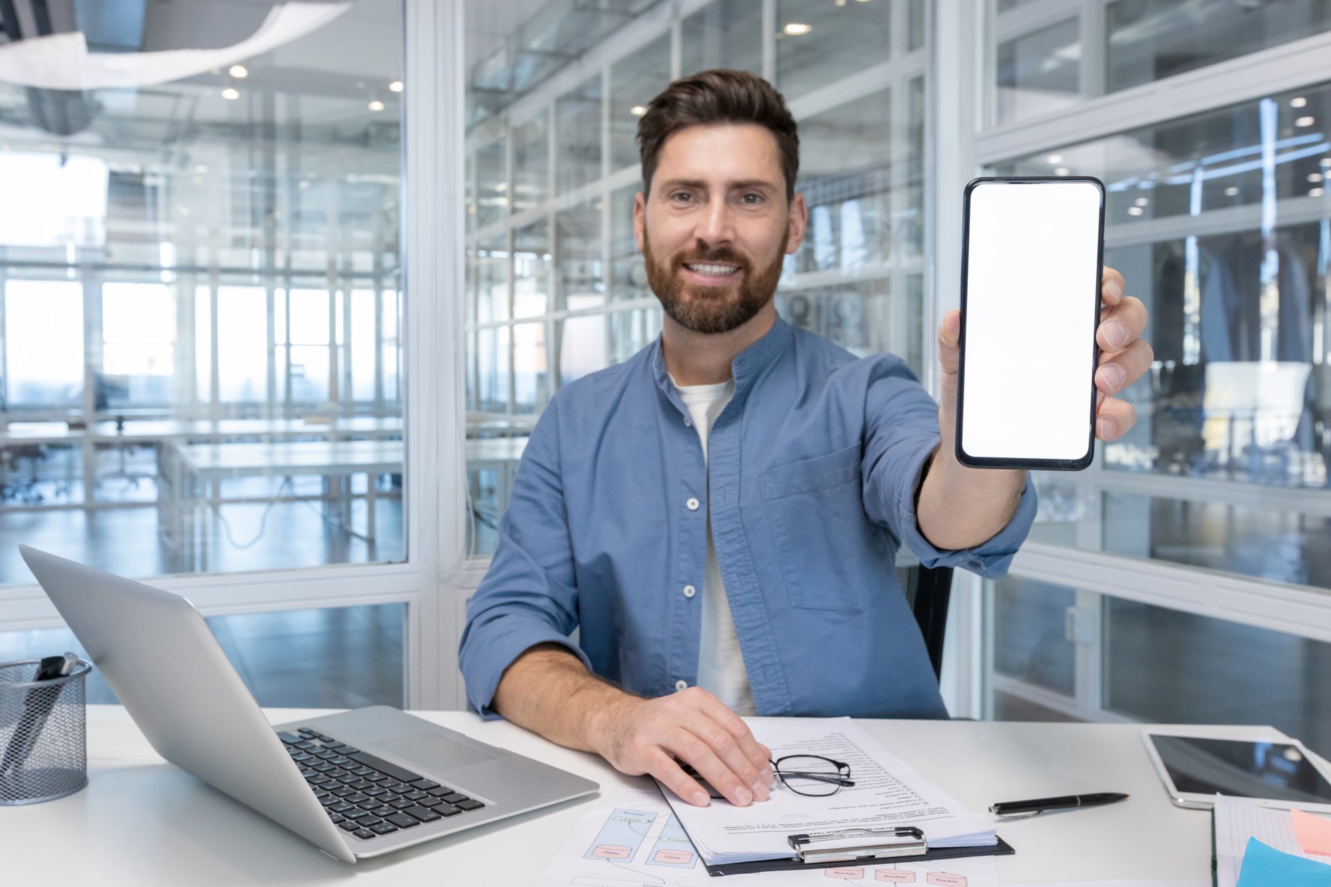 A cheerful man in a blue shirt displays a blank smartphone screen, smiling in an office setting with a laptop and documents.