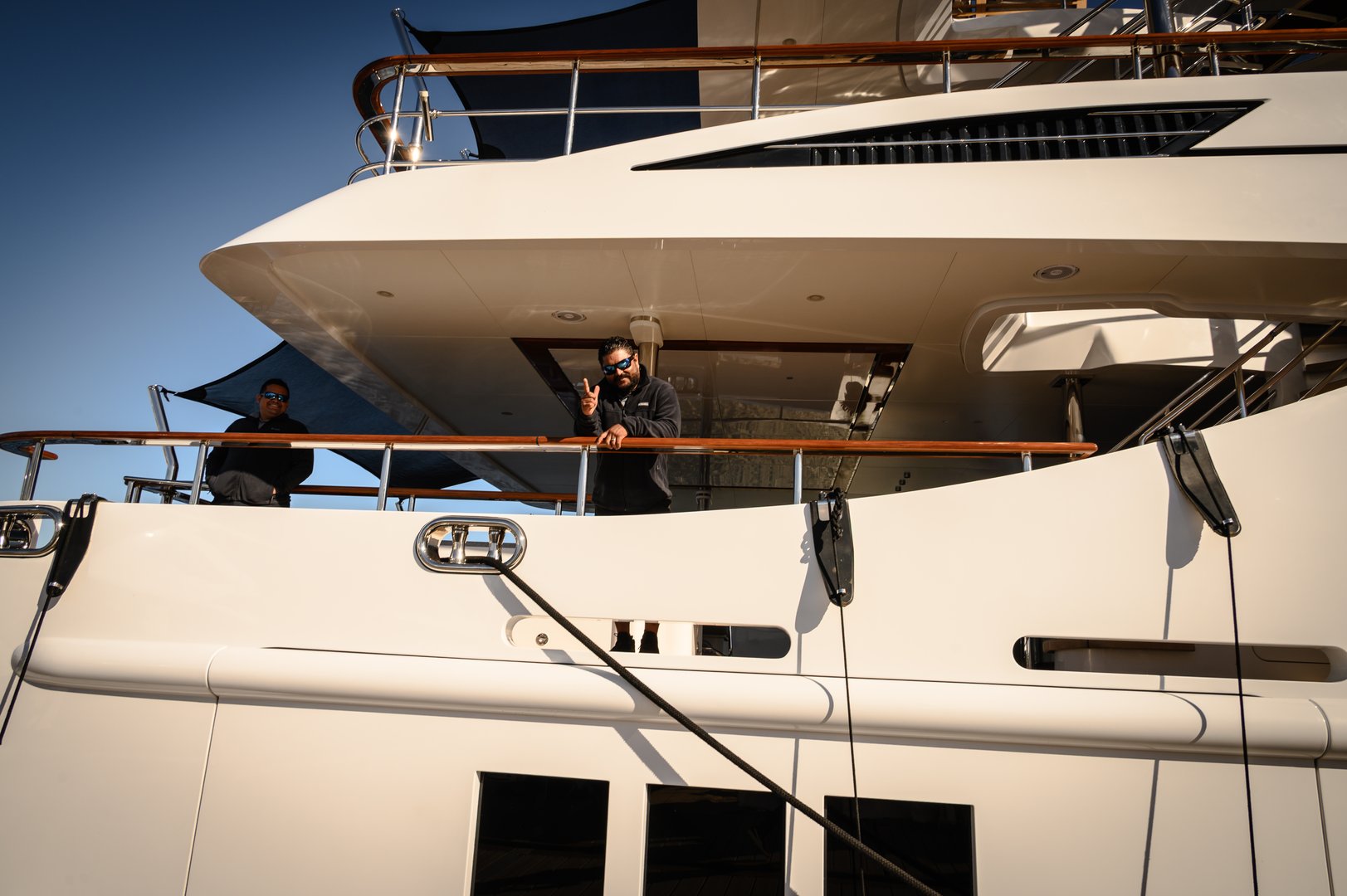 Two men stand on the upper deck of a sleek white luxury yacht during Fort Lauderdale's boat show, gazing toward the camera with one giving a friendly gesture. The scene highlights premium design, outdoor deck space, and a sunny, clear sky, conveying wealth, leisure travel, and maritime lifestyle.